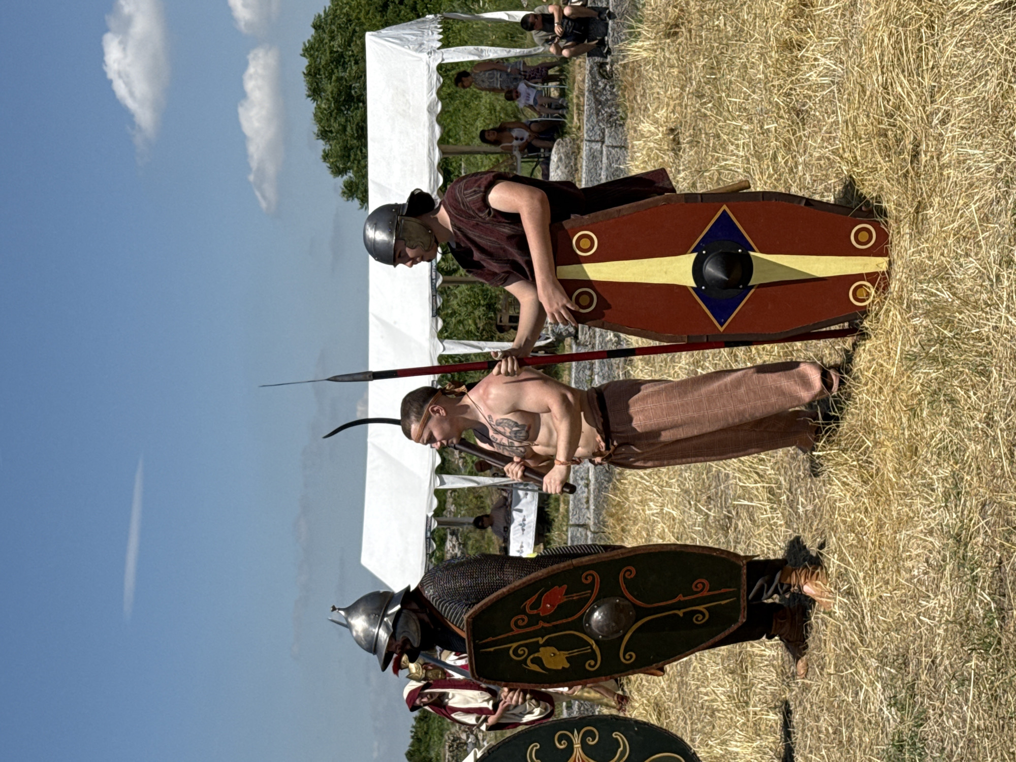 The image depicts a historical reenactment scene outdoors in the city of Gigen, Bulgaria, under a clear blue sky with sparse clouds. The foreground features three individuals dressed in ancient military attire, standing on dry, sparse grass.
From left to right:
1. A person, partially visible, wearing a pointed silver helmet and chainmail armor, holds an oval-shaped dark green shield adorned with yellow and red bird and spiral motifs, and a central metal boss. Behind this individual, a figure in a white and red garment holds a white shield.
2. A bare-chested individual, likely portraying a Dacian or Thracian warrior, wears light brown patterned trousers and a brown headband. This person has visible tattoos on their chest and left arm. They hold a short, curved weapon resembling a falx in their right hand and a long spear with a dark, pointed tip in their left hand, resting on the shoulder.
3. A person in a dark red tunic and a silver Roman galea-style helmet holds a long, dark spear vertically in their left hand and a reddish-brown, semi-rectangular shield (scutum) in their right hand. The shield features a central yellow vertical stripe, a blue diamond enclosing a black boss, and two yellow circles at the bottom.
In the mid-ground, a white canopy tent is set up, under which several individuals are seated or standing, observing the scene. To the right of the tent, a woman stands with a child, and a man is seated on a low stone structure. Green trees and bushes are visible in the background, behind the tent. The ground is covered with dry, yellowish grass and some visible earth.