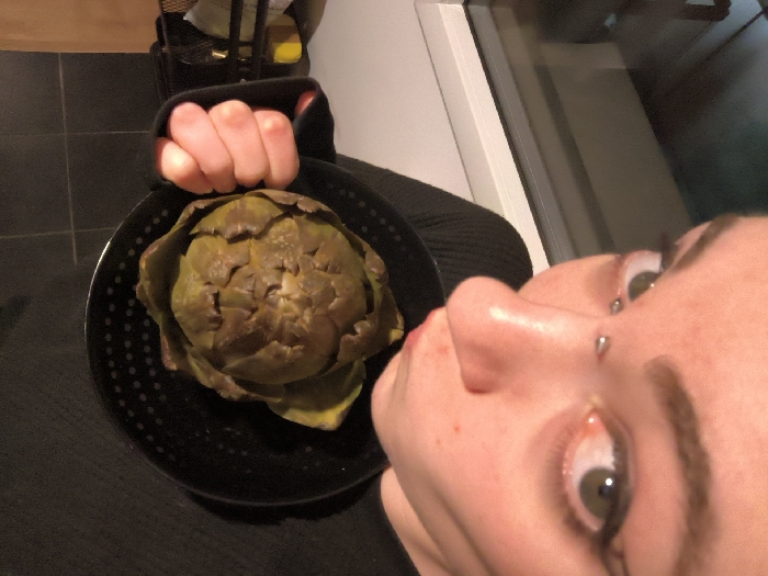 A close-up, angled shot focuses on a plate holding an artichoke. The plate is black with a speckled pattern. A hand grips the plate's edge. The artichoke is green and brown. Part of a person's face, including their nose, eyes, and eyebrows, is visible to the right of the plate, suggesting they are holding it. The background includes tiled flooring and a partially visible cabinet. The lighting appears to be indoor and possibly artificial.