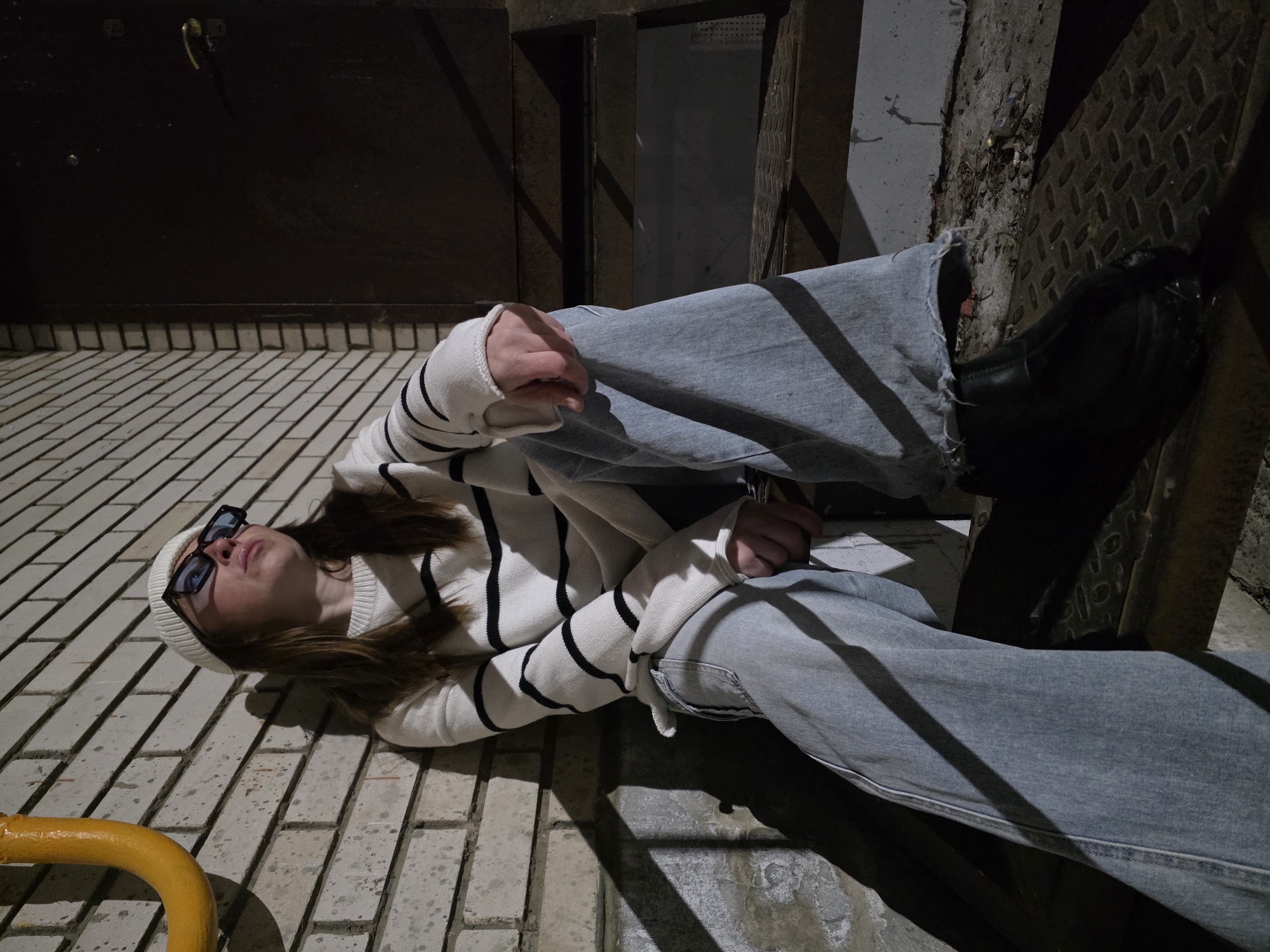 A female individual is depicted seated on dark metal steps, leaning against a light-colored brick wall in the city of Chaikovsky, Russia. The specific location within the city is not identifiable from the visual content. The individual wears a white knit beanie, black rectangular sunglasses, a white long-sleeved sweater with black horizontal stripes, light blue wide-leg jeans with frayed hems, and black shoes. Her gaze is directed upwards and to the right. Her left hand rests on her left thigh. A bright yellow, curved metal pipe is affixed to the brick wall in the upper left of the frame. To the right, a dark brown door with a brass-colored handle is partially visible. The metal steps, constructed from a textured or diamond plate pattern, appear worn and descend downwards. The ground adjacent to the steps shows some scattered debris. The scene is lit by a prominent light source from the left, casting strong, defined shadows onto the wall and steps.