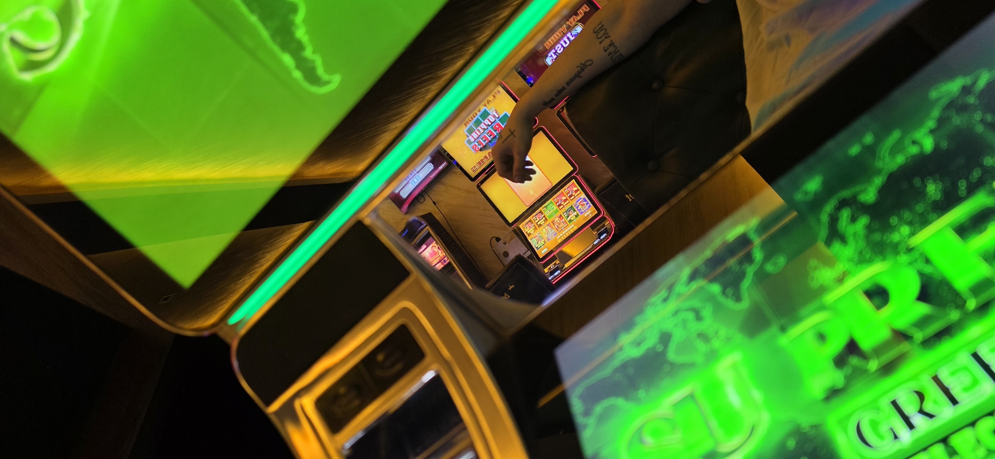 A person is playing a slot machine inside an establishment, possibly a casino, in Smyadovo, Bulgaria. The slot machine has multiple screens displaying colorful graphics and a hand is touching one of the screens. The surrounding environment is illuminated by neon green lighting. The person is seated on a dark, cushioned chair and has tattoos on their forearm.