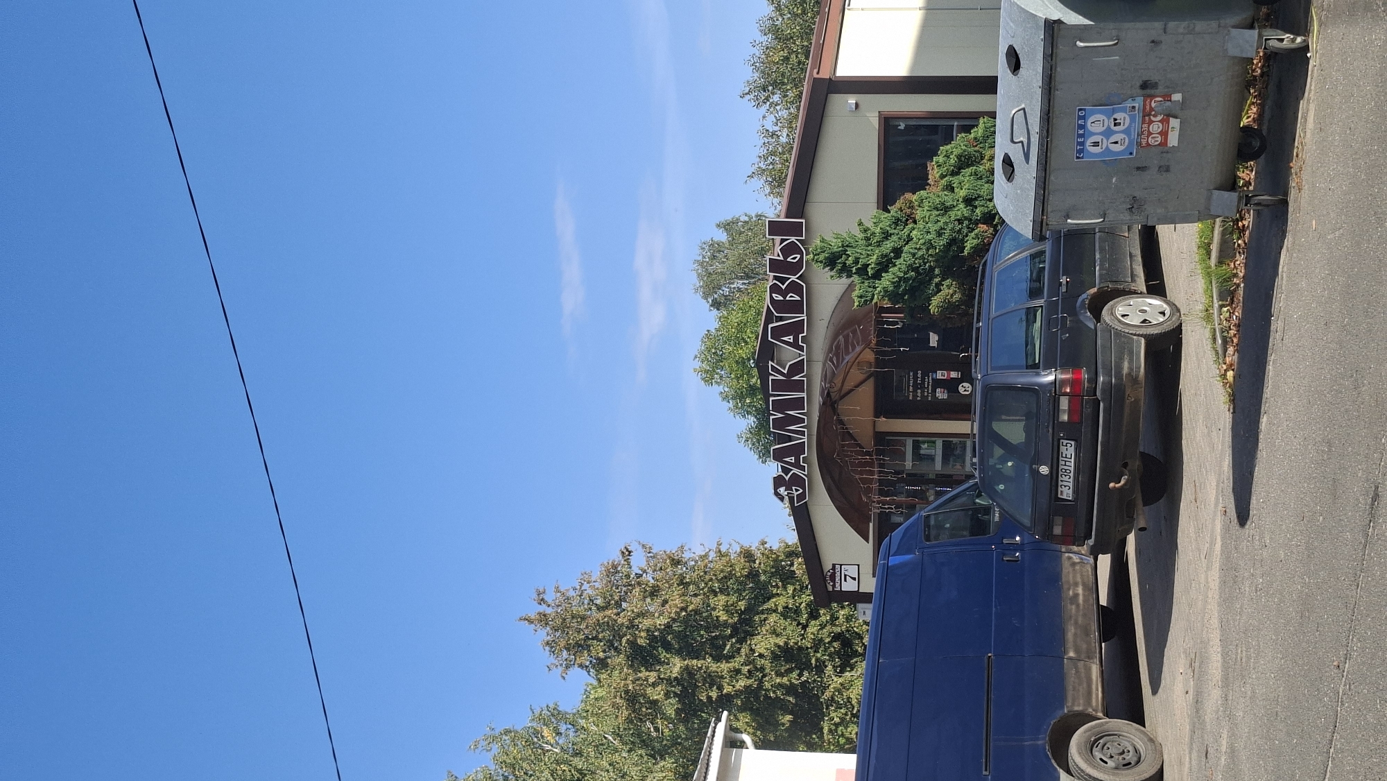 The image shows a street scene with a building and two parked cars. The building has a sign that reads "ЗАМКАВЫ" in large letters. Below the sign, there is an entrance with lights strung across it, and a smaller sign that displays business hours from 9:00 to 21:00. A number "7" is visible on a sign to the left of the entrance. To the left of the building is a white structure with a gutter.
In front of the building, two cars are parked on a paved surface. The car closer to the viewer is a dark-colored station wagon with a license plate that reads "3138 HE-5". To the left of this car is a blue van. A grey dumpster with a smiley face drawn on it is positioned to the right of the station wagon. The scene is set against a clear blue sky, with trees visible in the background. The overall impression is a commercial establishment in a moderately urban or suburban environment in Myadzyel, Belarus.