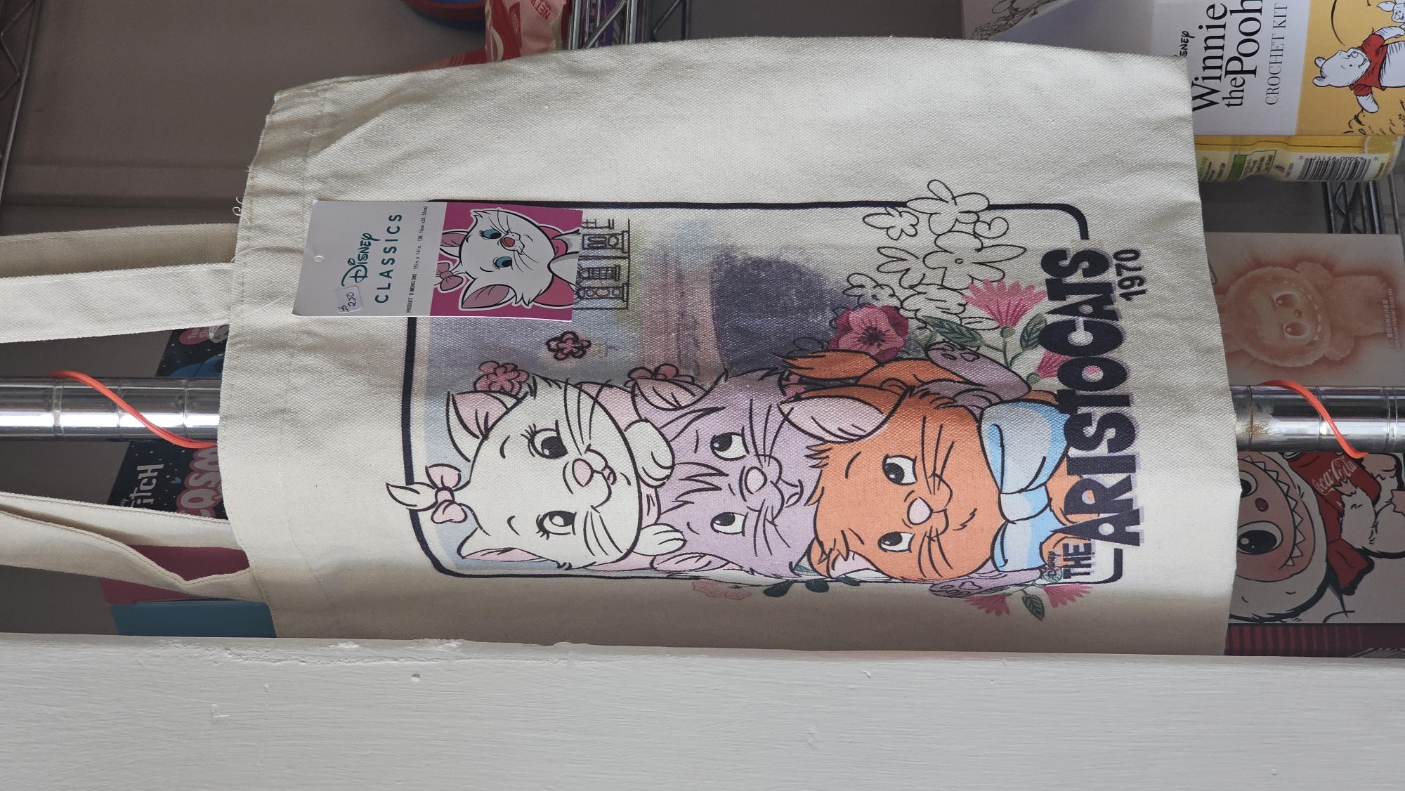 A close-up shot shows a white tote bag with a print featuring characters from Disney's "The Aristocats" film, placed on a shelf, seemingly within a residential setting. The tote bag is a light, off-white color and features a graphic design of the three kittens - Marie, Berlioz, and Toulouse - along with floral elements and the title "The Aristocats 1970." A Disney Classics tag is attached to one of the tote bag's handles. The shelf also holds other items, including a "Winnie the Pooh" crochet kit and various other objects that are partially obscured. Two horizontal metal poles run through the bag's handles. The location appears to be indoors. The image does not provide enough information to determine whether the setting is in the city of San Bernardino Tlaxcalancingo, Mexico.