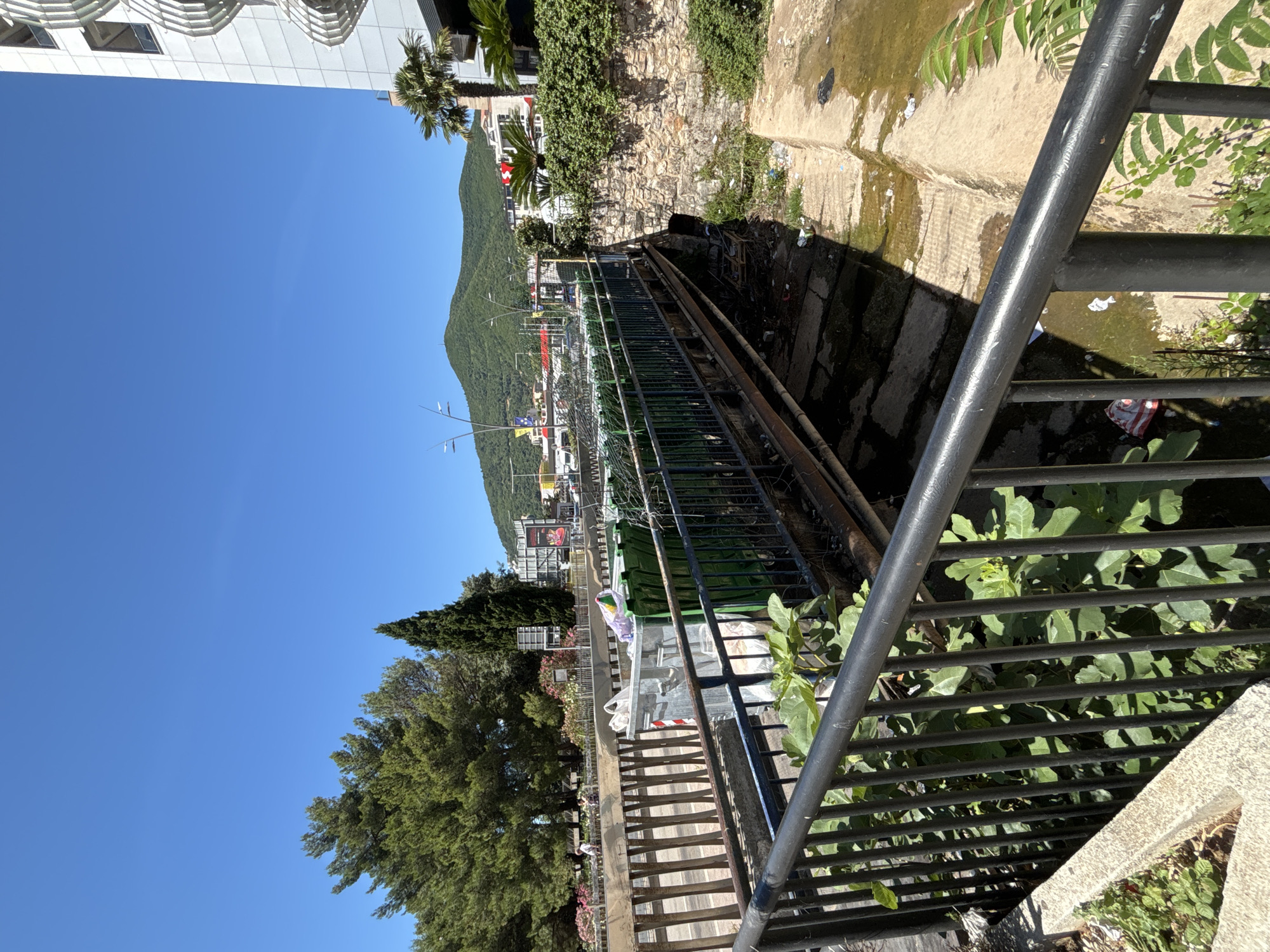 The image captures a scene in Budva, Montenegro, focusing on a bridge over a dry or low-water creek bed. The bridge, constructed of metal beams and railings, is lined with numerous green trash bins, some of which appear to be overflowing with garbage bags. A silver dumpster is also present on the bridge. The creek bed itself is littered with various debris, including plastic bags and other refuse.
The surrounding environment includes lush green trees, a stone wall with an arched opening under the bridge, and a modern white building with a distinctive wavy facade on the right side of the frame. In the background, rolling green hills are visible under a clear blue sky. A black metal railing is in the foreground, obscuring some of the lower portion of the scene, and green foliage is growing near it. The overall impression is one of a somewhat neglected urban area with signs of waste accumulation.