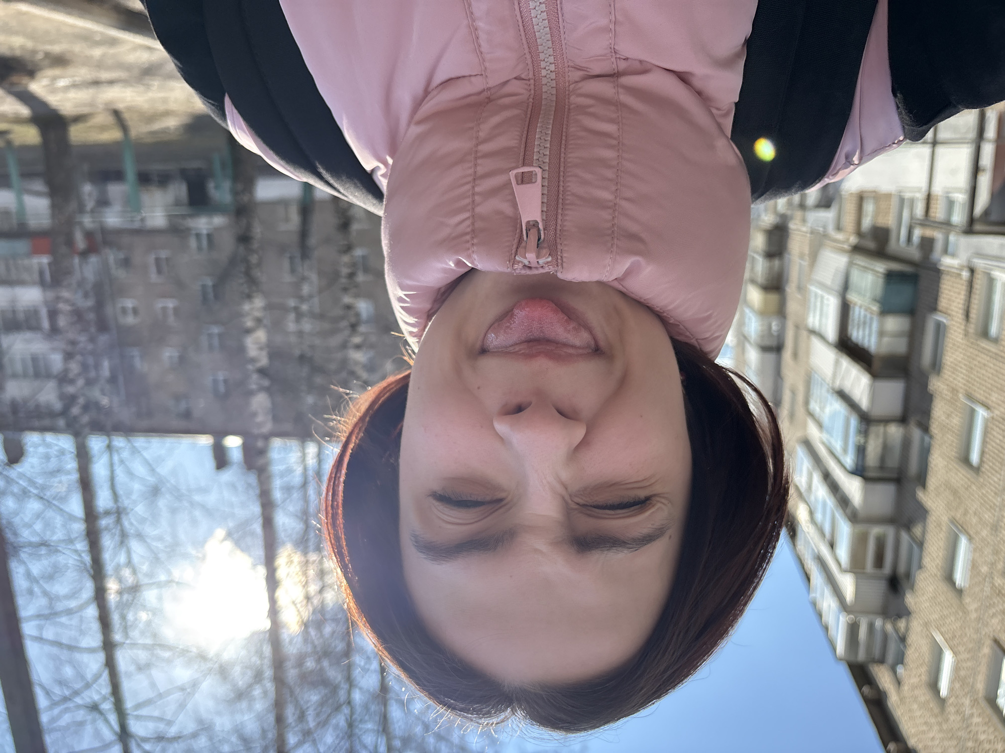 The image features a close-up, upside-down portrait of a person making a playful face outdoors. The individual, likely female, occupies the lower and central part of the frame (when viewed in its current orientation). Her eyes are squinted tightly shut, and her tongue is extended, suggesting a silly or mischievous expression.
She is wearing a pink puffer jacket with a prominent light-colored zipper running down the center. Dark-colored straps, possibly from a backpack or another garment, are visible over her shoulders.
The background, also inverted, depicts an urban or suburban environment. On the left side, bare tree branches are silhouetted against a bright, hazy sky with a strong sun glare. Beyond the trees, the faint outlines of residential buildings can be seen. On the right side of the frame, a multi-story brick building with numerous windows and balconies is clearly visible, characteristic of residential architecture. The bright, clear sky indicates daytime, and the bare trees suggest a season like late autumn, winter, or early spring in Chaikovsky, Russia.
The activity captured is a candid, playful pose for a photograph. No visible text is present in the image.