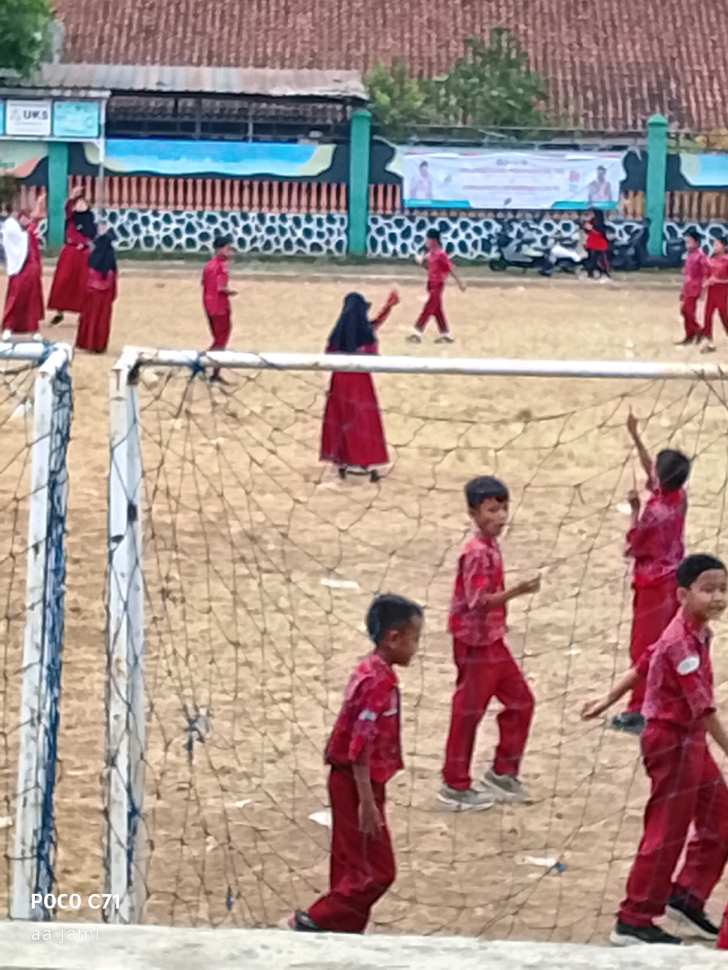 The image depicts a group of schoolchildren in red uniforms participating in an outdoor activity on a dirt field, possibly a sports event or physical education class. The setting appears to be a schoolyard, with a building and a fence in the background. A soccer net is prominent in the foreground, suggesting a game or practice is underway. Some children are actively engaged, while others appear to be observing or in transit. The overall scene conveys a sense of youthful energy and communal gathering.