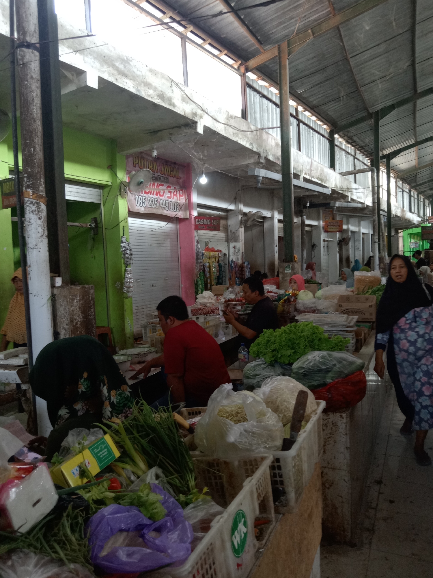 The image depicts a bustling outdoor market scene, likely in Sugihwaras, Indonesia. A corrugated metal roof supported by metal beams covers the stalls, with open sides allowing natural light to filter in. The foreground is dominated by various produce displayed in plastic bags and baskets. Among the visible items are bunches of green onions, leafy green vegetables such as lettuce, and what appear to be noodles or shredded ingredients in plastic packaging.
Several individuals are present. A woman in a black hijab and a patterned dress is walking towards the right side of the frame, her face partially obscured by shadow. In the middle ground, a person wearing a red shirt is seated, appearing to engage with a transaction. To their left, another individual is also seated, with their back to the camera, wearing dark clothing with a floral pattern. Further back, more people can be seen browsing or working at stalls.
The market appears to be well-stocked, with a variety of goods available. Signs are visible on some of the stalls, though the text is largely illegible. The overall atmosphere suggests a typical day of commerce in a local Indonesian market. The lighting indicates it is daytime.