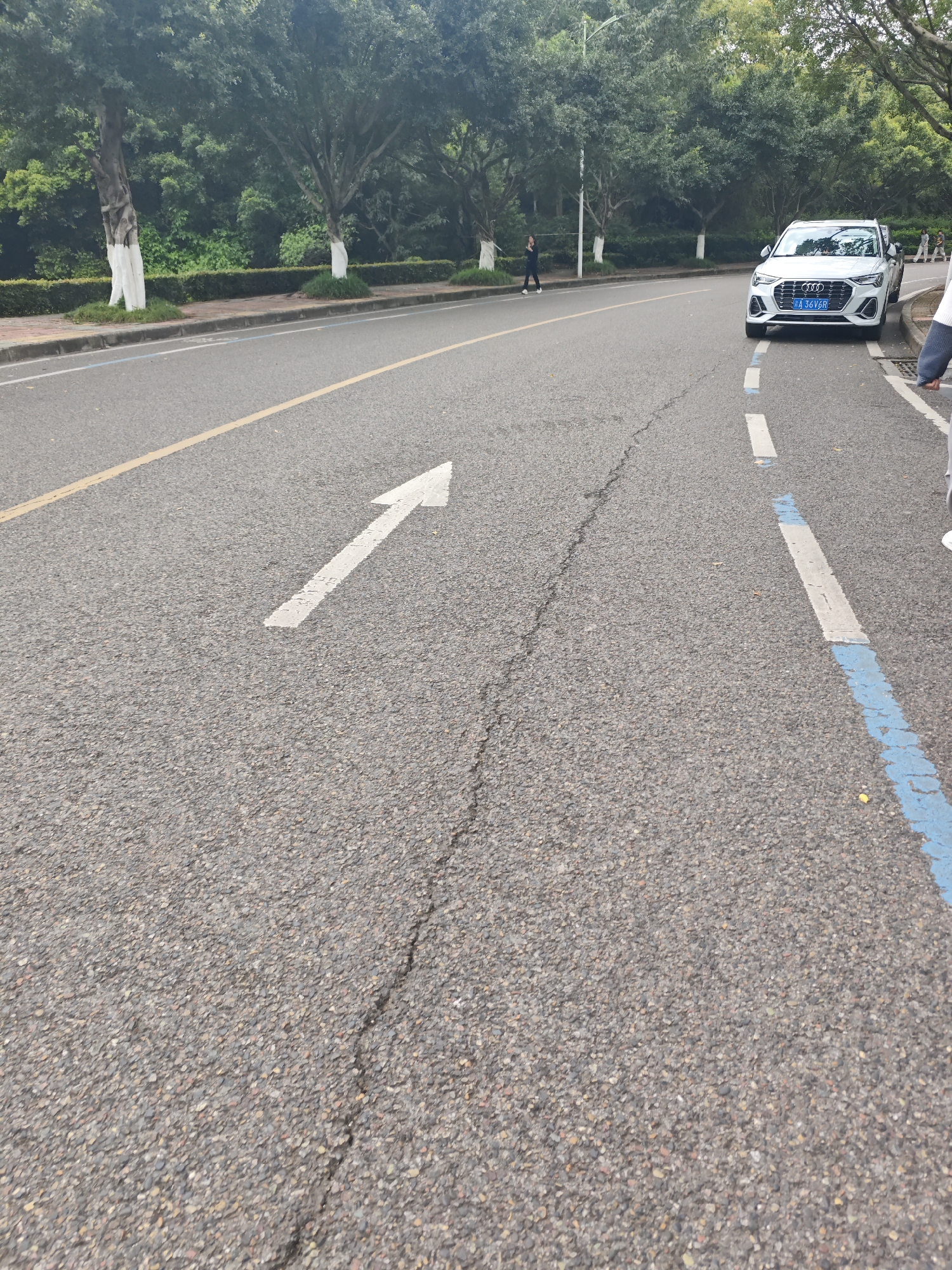 The image shows a paved road in Chongqing High-tech Zone, China. A white Audi with license plate "闽 A 36V68" is parked on the right side. A pedestrian walks on the left side of the road. A white arrow is painted on the road surface, directing traffic to the left. A yellow line marks the center of the road, and a blue-and-white line marks the parking lane. Trees line the side of the road. A crack runs down the center of the foreground. A person is standing on the right side of the frame.