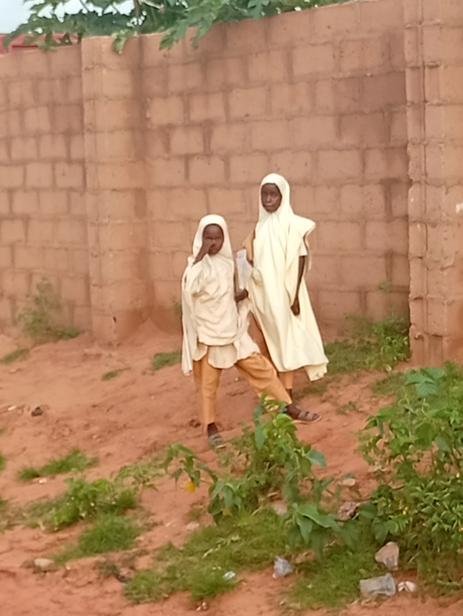 Two young people, appearing to be children, are standing in front of a brick wall in Tumfure, Nigeria. Both are wearing light-colored, loose-fitting garments and head coverings. The individual on the left is positioned slightly behind and to the left of the person on the right. The ground is covered in reddish-brown dirt with patches of green vegetation. The brick wall behind them is constructed from rectangular, light brown bricks and appears to be part of a building or enclosure. The overall lighting suggests it is daytime.