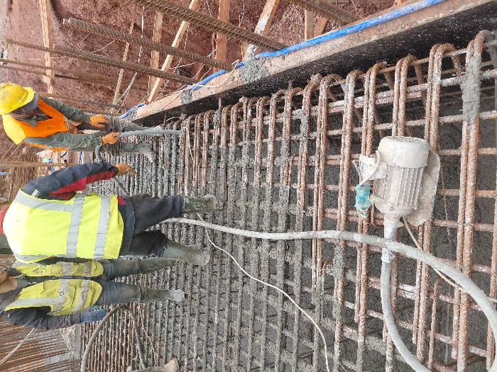 The image depicts a construction site in Fdalate, Morocco, showing concrete pouring activity for a structural element. Three male construction workers are present on a grid of steel rebar partially submerged in fresh grey concrete.
On the left, one worker (partially visible) wears a yellow high-visibility vest over a dark jacket, blue gloves, dark pants, and green rubber boots. In the center, a second worker stands with his back mostly to the camera, wearing a yellow high-visibility vest over a dark and red long-sleeved shirt, dark pants, and green rubber boots. On the right, a third worker, wearing an orange high-visibility vest, a yellow hard hat, orange gloves, and green rubber boots, is actively using a concrete vibrator (a tube-like device) to consolidate the wet concrete.
The work surface consists of a dense, rectangular grid of rust-colored steel rebar. Wet grey concrete covers a significant portion of this rebar grid. A flexible, grey concrete delivery hose, partially encrusted with concrete, traverses the rebar from left to right. An inactive, dusty beige electric motor with a green cap and an attached flexible shaft lies on the rebar grid in the foreground. Along the right edge of the pour, dark wooden formwork (shuttering) contains the concrete, with blue conduits or pipes attached to its side. Beyond the formwork, an excavated area of reddish-brown earth and additional rebar structures or timber supports are visible. The overall environment suggests an outdoor construction setting.
