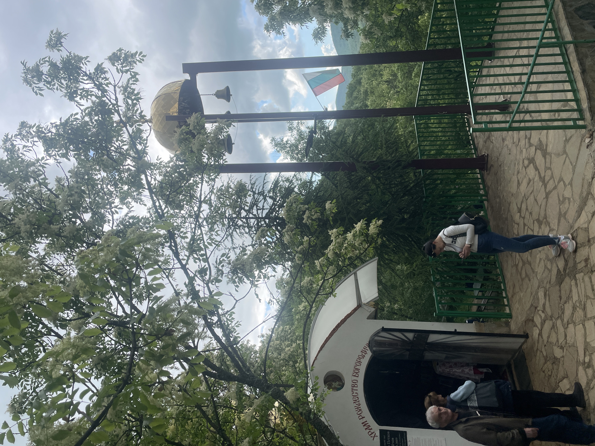 The image captures a scene at the Monastery "Rozhdestvo Bogorodichno" in Novo Selo, Bulgaria. In the foreground, several people are standing near a small, white building. One woman, dressed in jeans and a white top, is looking at her phone. Two more people are entering the building.
Above, a golden dome and bells are visible, positioned over a green metal fence. In the background, a Bulgarian flag is flying. The environment is characterized by lush greenery and trees. The sky is partly cloudy.