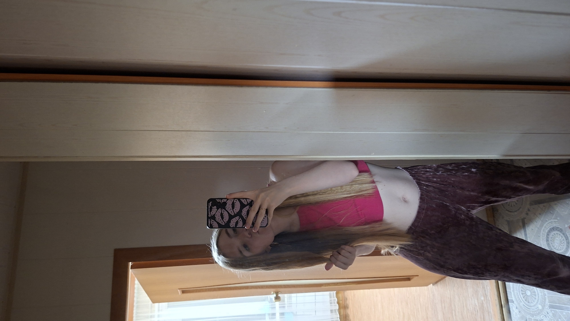 A young woman is captured in a mirror selfie, holding a smartphone to photograph her reflection. The image is oriented sideways, but if viewed upright, shows her from roughly the waist up. She has long, straight blonde hair and is wearing a bright pink sleeveless crop top that exposes her midriff, paired with loose, dark-colored pants, which appear to be made of velvet or a similar textured fabric. Her expression is neutral as she focuses on the phone.
The smartphone is held in her left hand, and its case is black with a repeating pattern of white or light pink lips/kisses. Her right hand is seen near her hair, possibly adjusting it.
The background, reflected in the mirror, consists primarily of light-colored wooden paneling, suggesting the mirror is part of a wardrobe or closet door. In the lower left of the upright image, a light-colored wooden door frame is visible, along with a reflection of what appears to be a window or light source. The flooring on the right side features a light-toned tile or linoleum with a subtle, circular geometric pattern. The ambient lighting indicates it is daytime. The scene suggests an indoor, residential setting, with the provided location context being Большой Букор, Russia. No legible text is visible within the image.