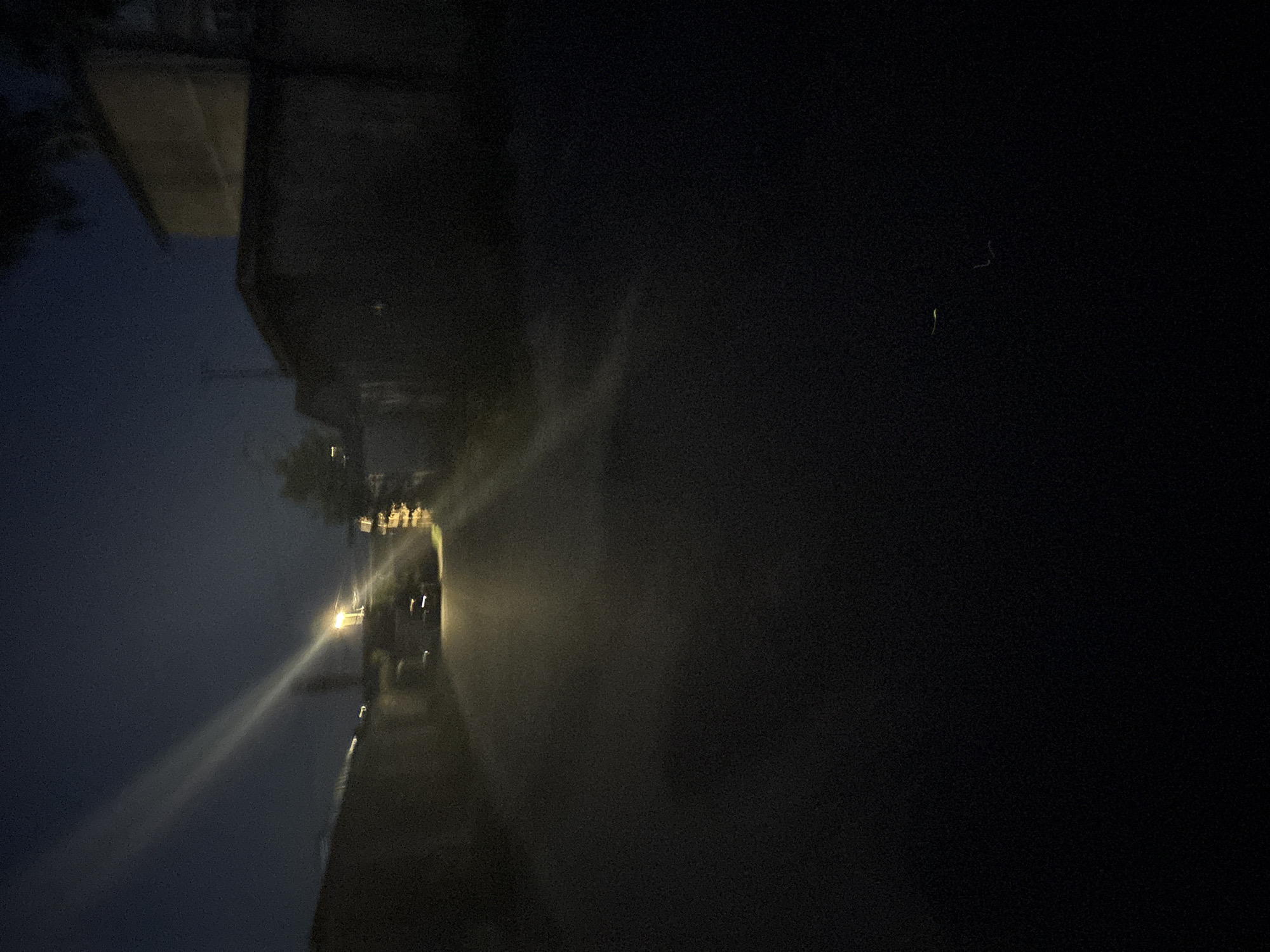 The image depicts a nighttime scene on a dark, likely paved road or alleyway that recedes into the distance. A prominent overhead streetlight, emitting a bright yellowish-white light, is positioned in the mid-ground, slightly right of the central axis. A distinct, elongated diagonal beam of light, consistent with a lens flare or atmospheric scattering, extends upwards and to the left from the streetlight. The sky above is a deep indigo or dark blue, indicative of night.
On the left side of the road, a dark, indistinct mass, possibly a wall, fence, or dense foliage, lines the path, with a lighter-colored structure barely visible further back. To the right, a large, dark, block-like building (possibly an industrial or commercial structure) occupies the immediate foreground, with the upper portion of a lighter-toned building visible behind it. Dark foliage, possibly tree branches, is partially visible on the far right edge.
Beneath the main streetlight, a dark vehicle, possibly a car, is partially illuminated and faces away from the viewer. Faint, small horizontal streaks of light in the distance suggest additional obscured light sources or vehicles further down the road. The foreground and lower portions of the image are predominantly dark, with significant digital noise or grain evident, particularly in these low-light areas. No individuals are discernible within the frame.
The specific location within the city of Unknown, Unknown, cannot be determined from the visual evidence.