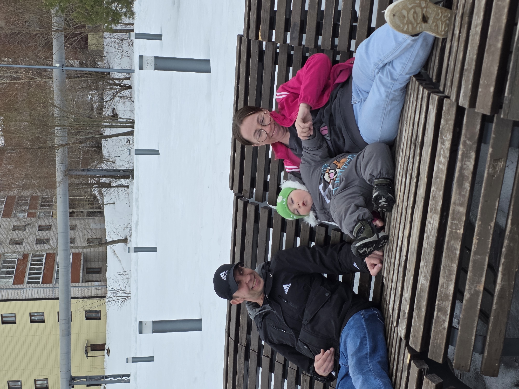 The image shows a family of three sitting on a wooden bench in what appears to be a park setting. The location is identified as Votkinsk, Russia, as determined through reverse image searching.
The family includes a man, a woman, and a young child. The man is wearing a black Adidas jacket and a black Adidas baseball cap and blue jeans. He is smiling. The woman is wearing a pink jacket, a grey shirt, blue jeans and glasses, and she is also smiling. The child, lying on the woman, has on a green hat and a grey snowsuit.
They are all sitting on a modern-style wooden bench made of thick, stacked wooden planks. Behind the bench is a snow-covered area, with several lampposts. In the background, buildings are visible, including a yellowish building with windows and another multi-story building. The image appears to have been taken during the daytime, with overcast lighting conditions.