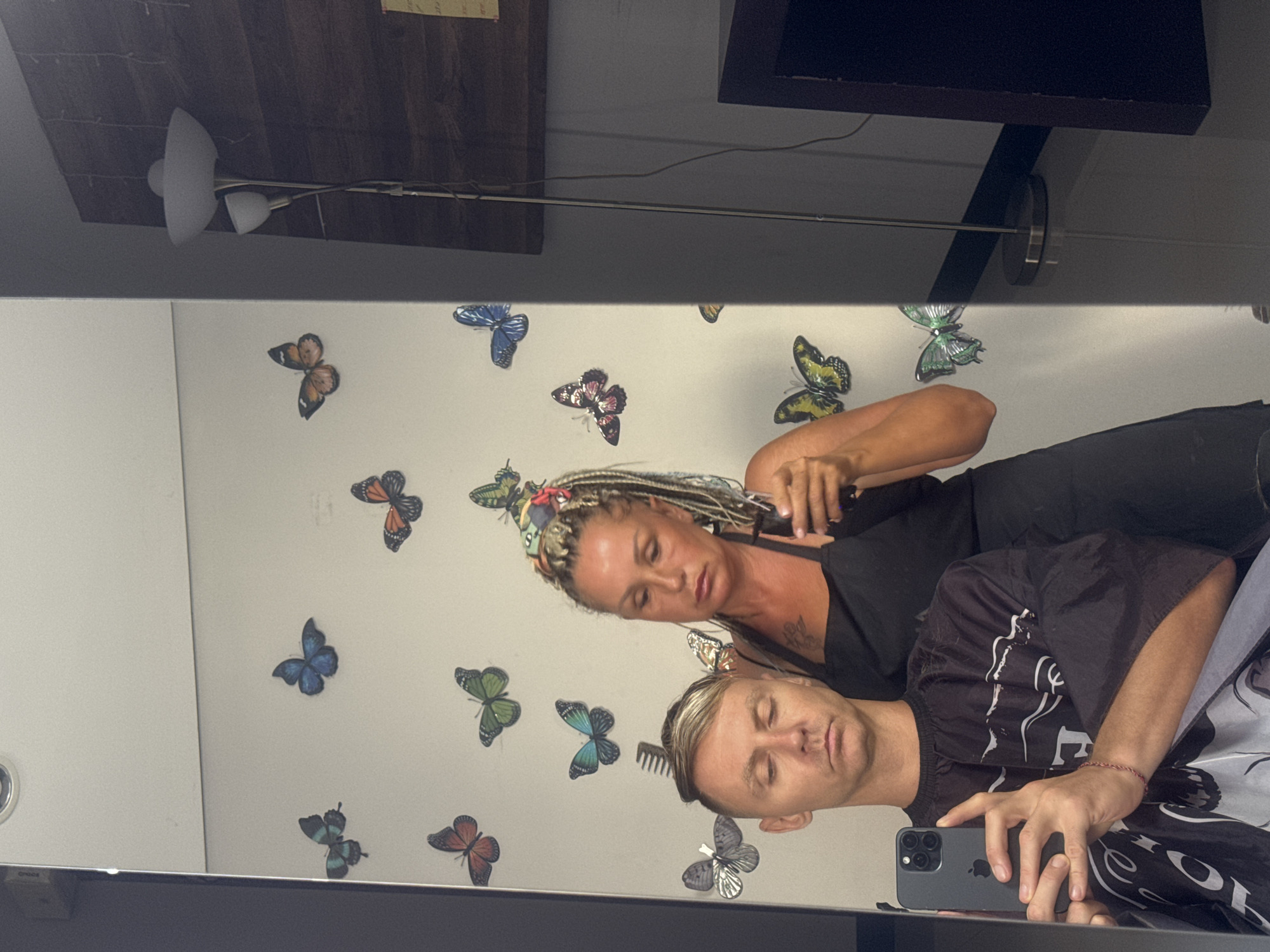 The image appears to be a selfie taken in a mirror reflection in what might be a hair salon. The reflection shows a woman, likely the hairstylist, with blonde braids and colorful headwear, wearing a black shirt, and tending to the hair of a man. The man, seemingly the person taking the selfie, is wearing a barber cape and holding an iPhone. The wall behind them is adorned with butterfly stickers. The environment includes a light fixture above, and a wooden shelf partially visible near the top of the image. There is no explicit indication of the location being Kuta, Indonesia, within the image itself.