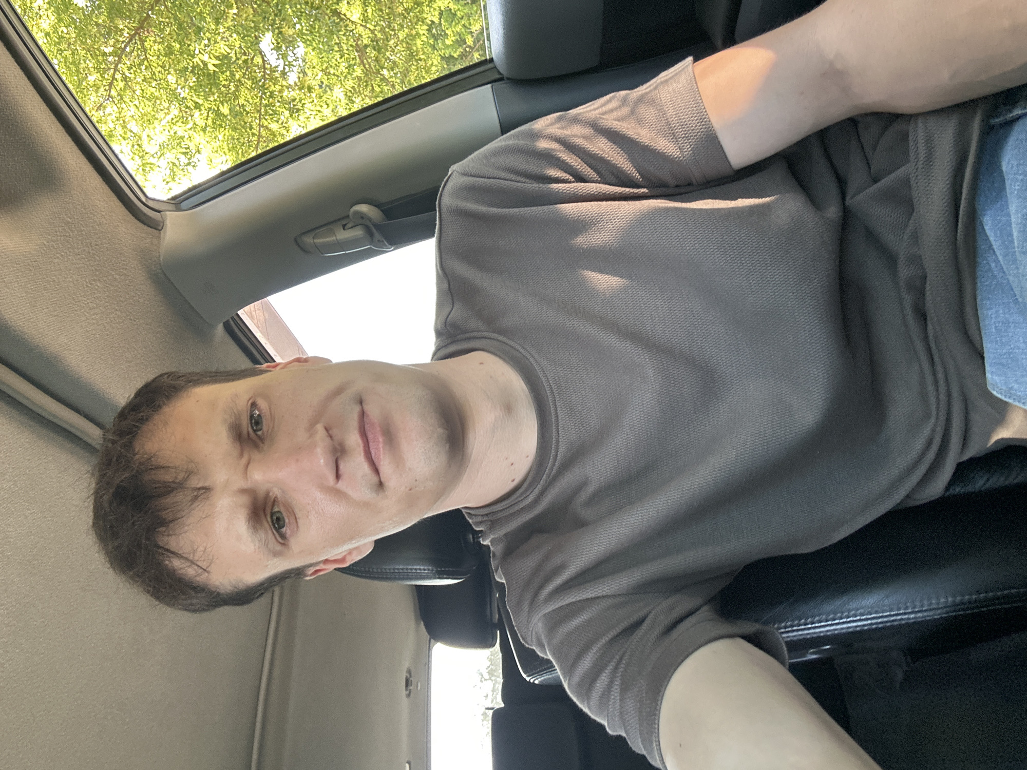 A medium shot captures a Caucasian man in his 30s sitting in the driver's seat of a car. He has light skin, brown hair styled neatly with a side part, and blue-green eyes. The man is wearing a plain, short-sleeved, brown t-shirt and blue jeans. Sunlight streams through the car's window, illuminating his right arm and shoulder. The interior of the car is visible, with black leather seats and the grey headliner of the car's roof. Outside the window, lush green trees are visible against a bright sky, suggesting it is daytime. The man is looking directly at the camera with a neutral expression. The photo appears to be a selfie taken inside a car, possibly in or near Makiv, Ukraine, given the context provided.