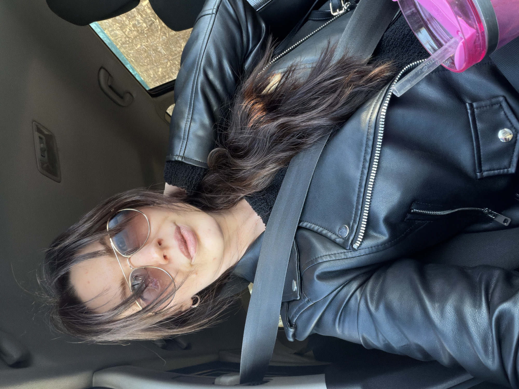 A woman wearing a black leather jacket, sunglasses, and a seatbelt is sitting in a car. Her dark brown hair is partially visible. A pink water bottle is positioned near her shoulder. The interior of the car and a partial view of what appears to be trees outside the window are visible. There is no evidence in the image to indicate the location is Perm, Russia.