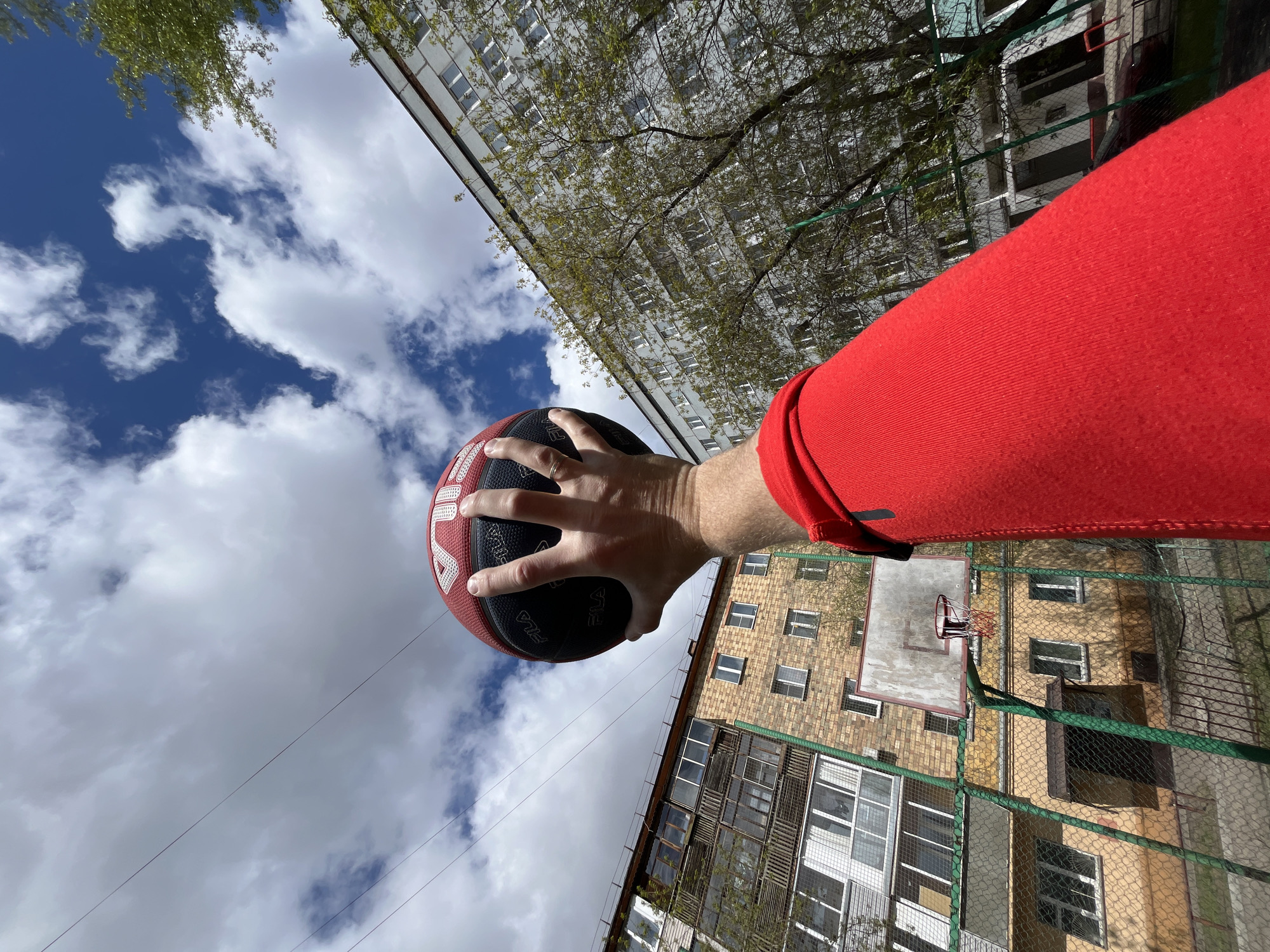 The image shows a person holding a Fila basketball in their left hand, wearing a red long-sleeved shirt. The backdrop features a cloudy sky and two buildings. One building is a high-rise apartment complex with numerous windows. The second building, partially obscured by a green chain-link fence, is of brick construction and appears to be a residential building. The location appears to be a basketball court in Krasnoyarsk, Russia. A basketball hoop is visible through the chain-link fence. Power lines are also visible against the cloudy sky.