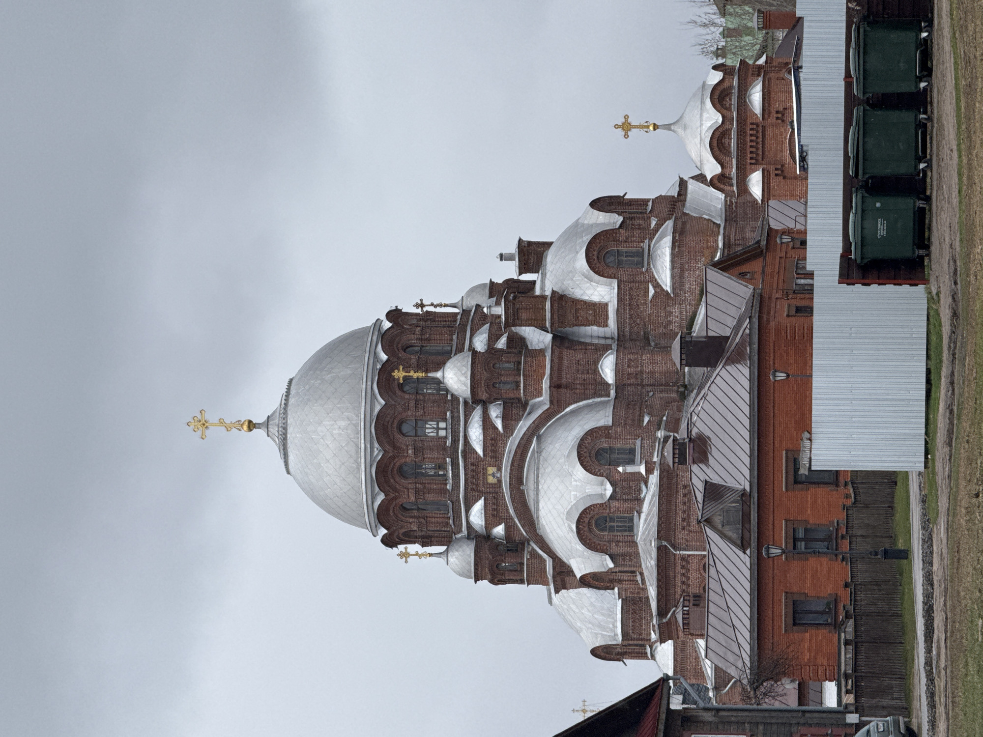 The image shows the exterior of the Cathedral of the Icon of the Mother of God of All Who Sorrow Joy. Located in Sviyazhsk, Russia, the brick building is characterized by its numerous silver domes, each topped with a golden cross. The architectural design includes arched windows, and the building appears to be several stories tall. To the right, there's a white structure with a corrugated metal roof, with green bins visible. The background is a gray sky. Parts of smaller wooden buildings are also visible in the foreground.