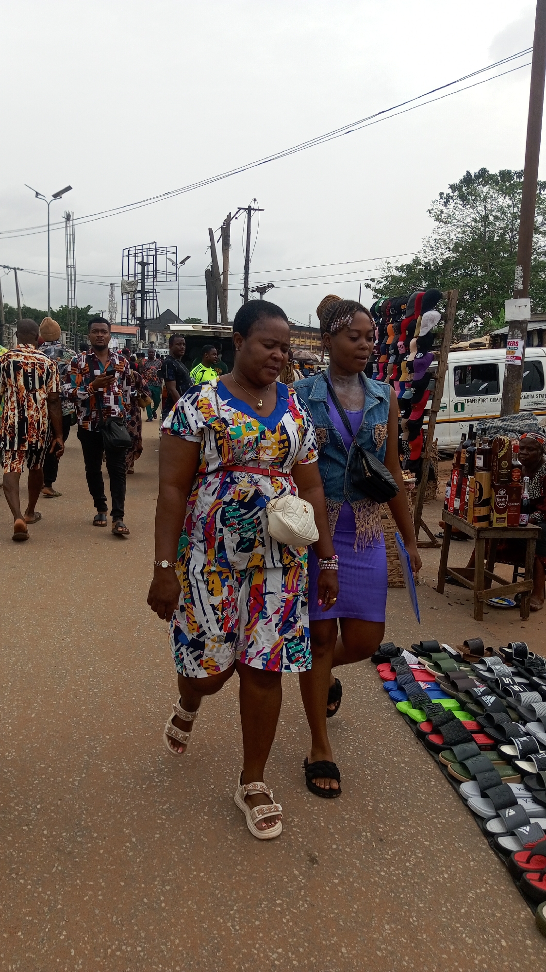 This image depicts a bustling outdoor market scene in Onitsha, Nigeria, under an overcast sky. In the foreground, two women walk towards the right. The woman on the left wears a colorful, abstract-patterned jumpsuit with a white background, accented by a red belt and a white quilted fanny pack. She also wears light-colored sandals, a gold necklace, and a watch. Beside her, the woman on the right is dressed in a purple sleeveless mini-dress, layered with a denim vest, and wears black slides. She carries a black cross-body bag and has braided hair styled upwards.
In the mid-ground and background, numerous other people are visible, moving through the market. One man in a patterned long-sleeved shirt is seen looking at his phone. On the right side of the frame, street vending activities are prominent. A large assortment of sandals and slides, some bearing visible brands like "ADIDAS" and "Y-3", are displayed directly on the reddish-brown ground. Further back, there's a rack of hats or caps. An elderly person is seated behind a small wooden table laden with various bottles of spirits, including "STAR", "HENNESSY", "NAPOLEON", and "REGAL". A white commercial van, partially visible, has text that reads "TRANSPORT COMM..." and "ANDRA STATE". The urban environment is characterized by utility poles, overhead wires, streetlights, and buildings, with a large, empty billboard structure in the distance. The day appears cloudy, with no harsh shadows.