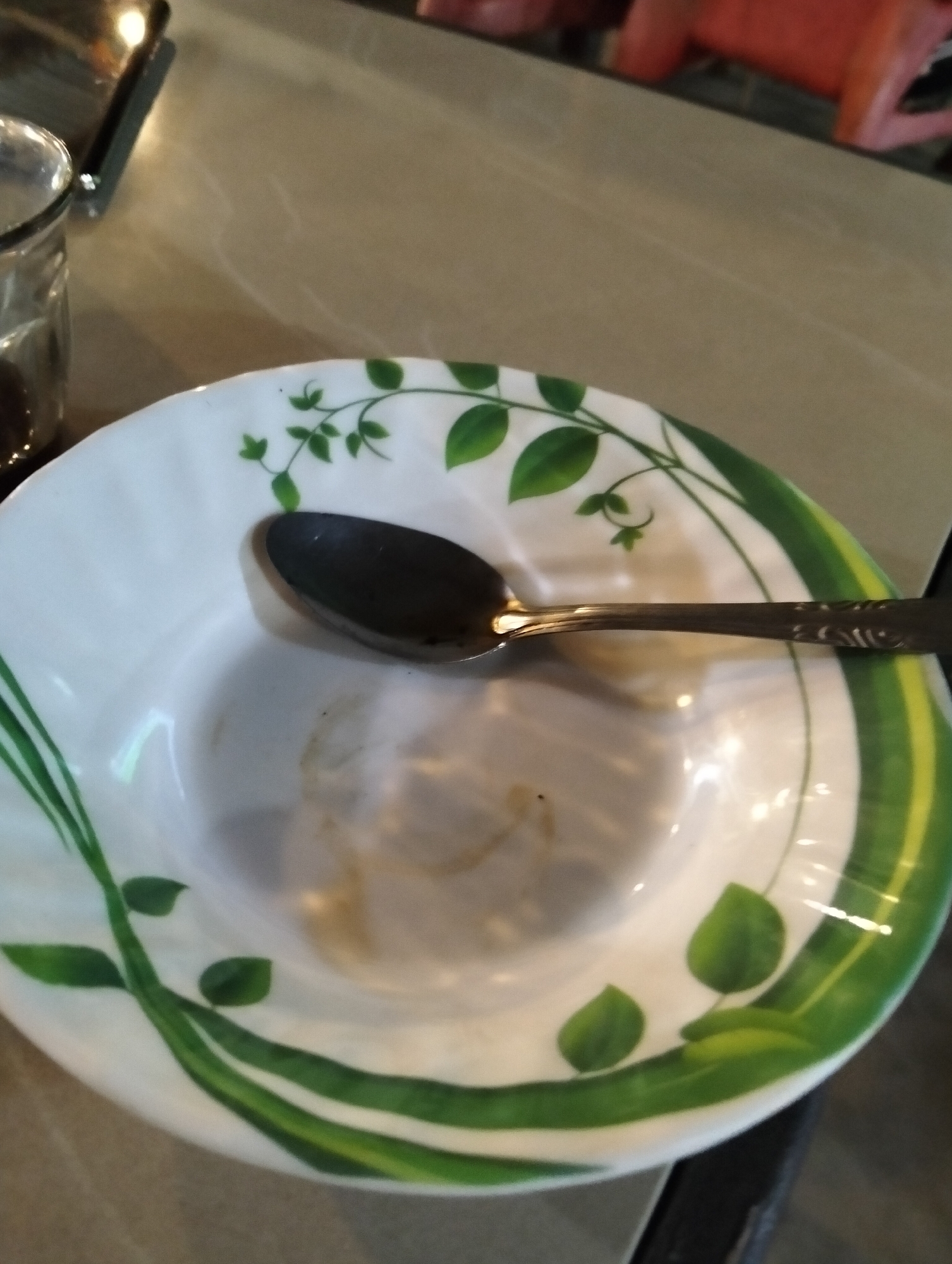 An overhead shot shows a white ceramic bowl with green vine and leaf decorations. A metal spoon rests inside the bowl, slightly off-center. The bowl contains some residual brown liquid or food remnants. To the left of the bowl, a glass with dark liquid is partially visible. The objects are placed on a table with a textured surface, reflecting light. The background shows a blurred reddish object and a dark surface. The scene appears to be indoors, likely in a dining setting.