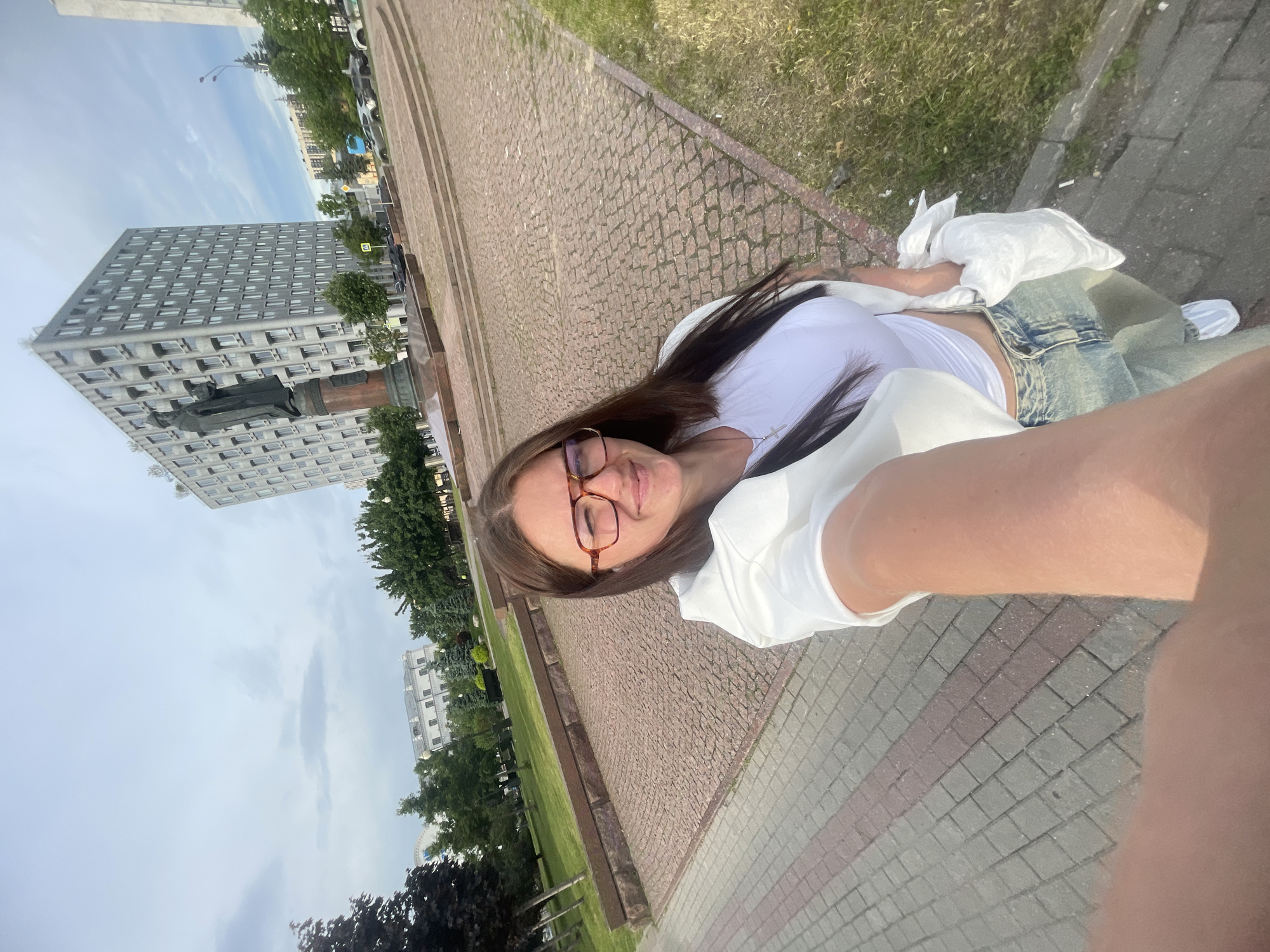 The image shows a young woman with brown hair and glasses taking a selfie in front of the House of Government of the Russian Federation in Moscow. She is wearing a white top and jeans. The background shows a park and other buildings. The weather appears overcast. The image appears to be taken at ground level from a low angle. The woman is partially sitting on an elevated, stone sidewalk.