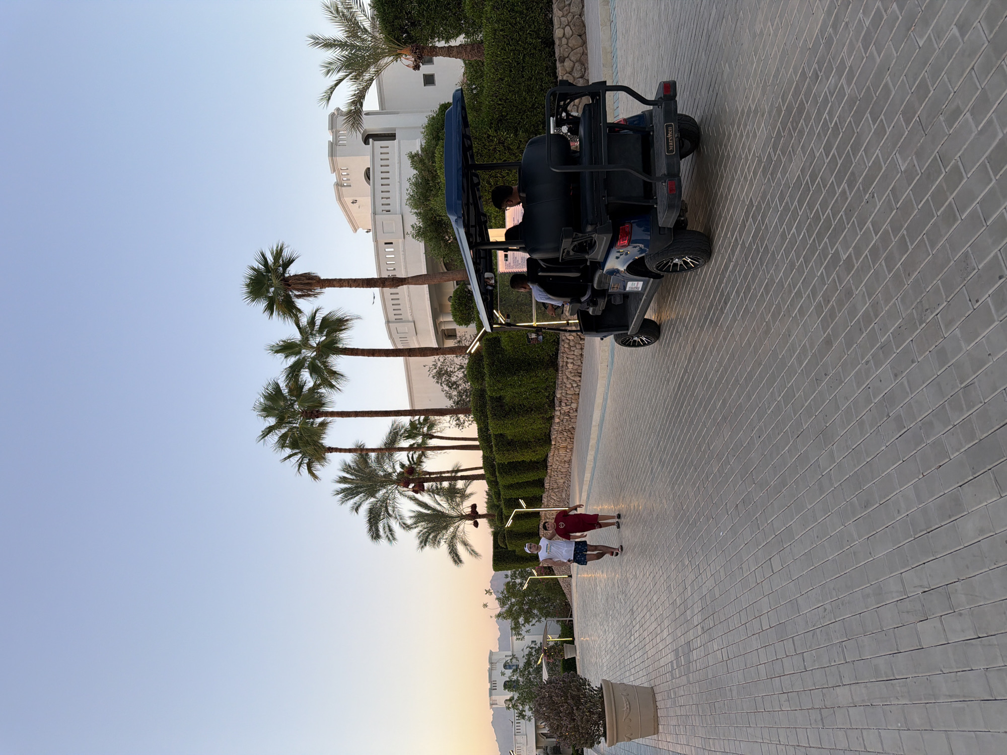 In Sharm El Sheikh, Egypt, two young boys walk down a paved walkway lined with palm trees and manicured hedges. A dark blue golf cart, with two individuals seated inside, travels in the same direction. In the background, a large white building with architectural details suggestive of a resort or hotel is visible, partially obscured by vegetation. The sky is clear and a soft sunset glow is present, casting a warm light on the scene. A large potted plant is situated to the left of the walkway. The overall environment suggests a well-maintained outdoor space, likely part of a tourist destination.