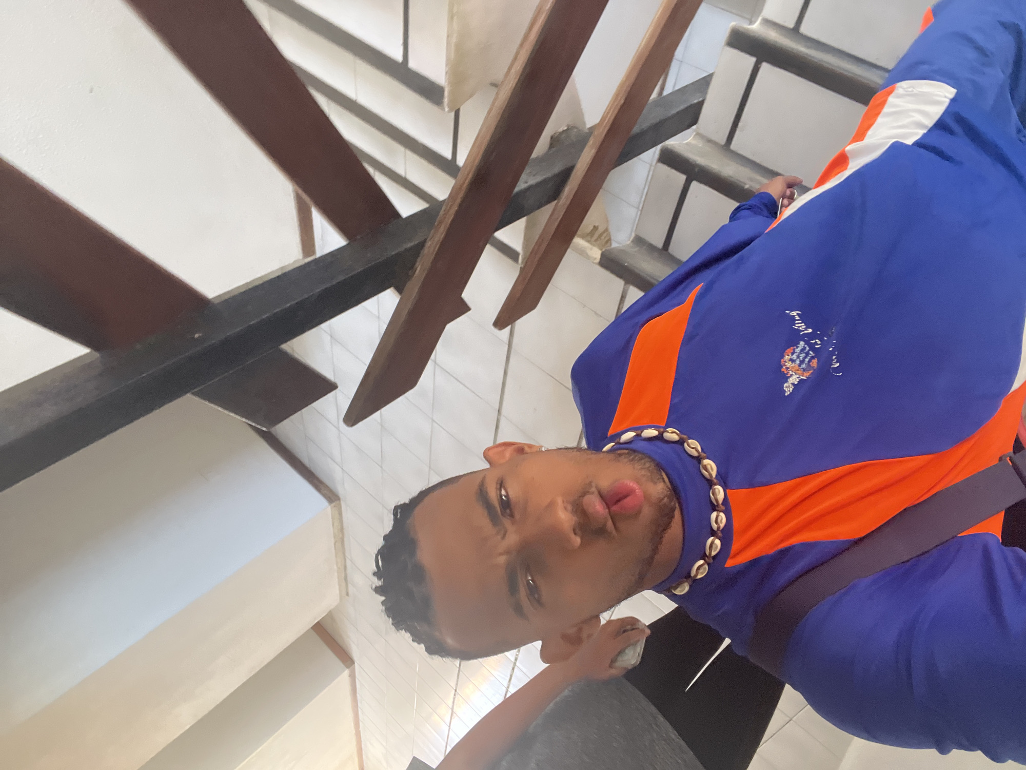 A medium shot, taken at an oblique angle, captures a young man standing on stairs. He is wearing a blue and orange jacket and a necklace made of seashells. The location is the city of Porlamar, Venezuela. The stairs have a wooden railing and a tiled surface. The man is making a pursed lip facial expression towards the camera. He is holding a small, unidentified white object in his left hand.