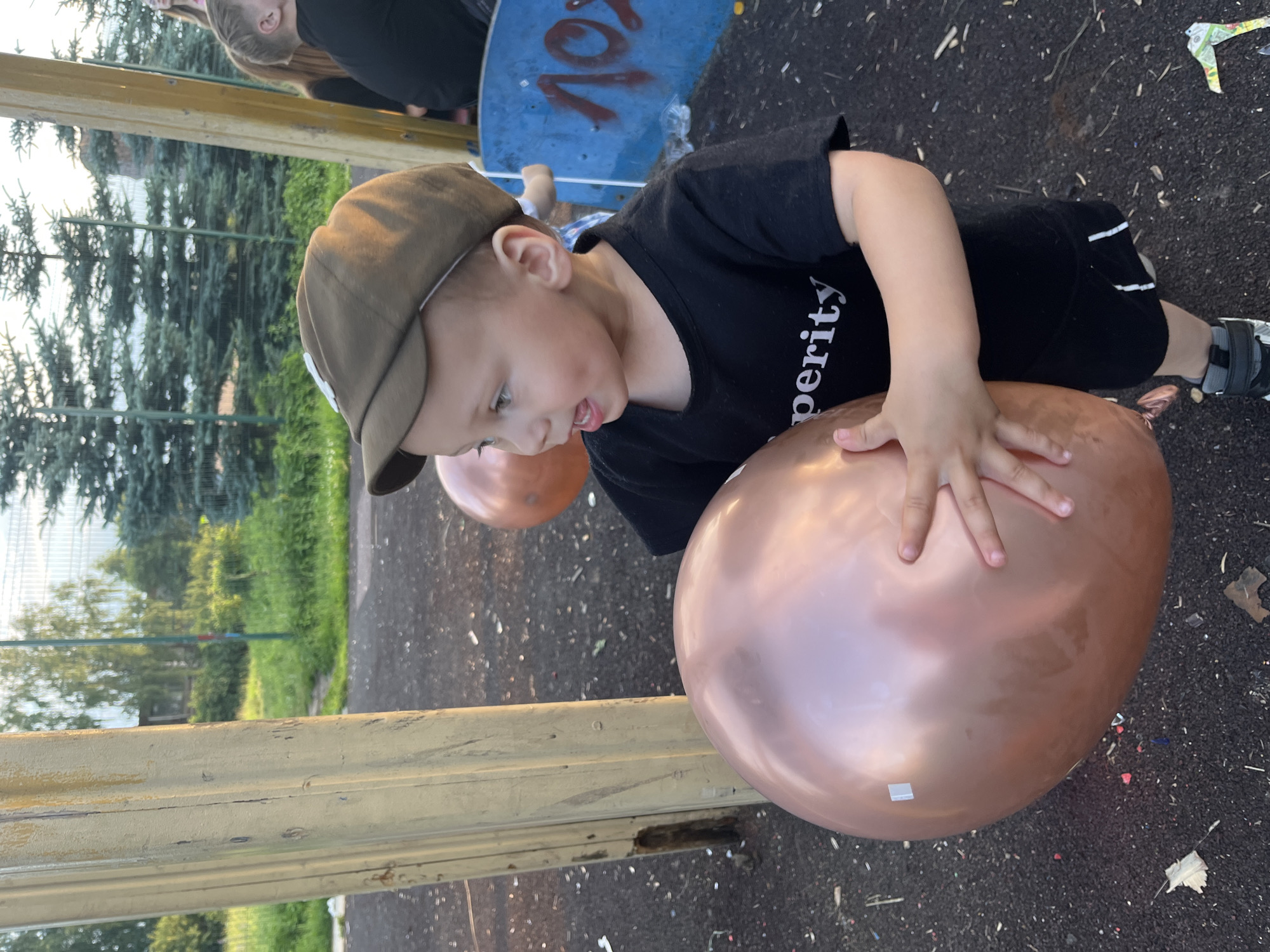 A young boy wearing a dark t-shirt with the word "perity" visible and a brown cap is holding a large, rose-gold balloon in an outdoor setting. He is looking to his left with his mouth open. Another balloon of a similar color is visible behind him. The ground is covered in dark, rubbery material, with some confetti scattered on it. A wooden structure is to the left of the boy. In the background, there are trees and a blue structure with some markings on it. The scene appears to be in a park or playground. The location is identified as Foki, Russia.