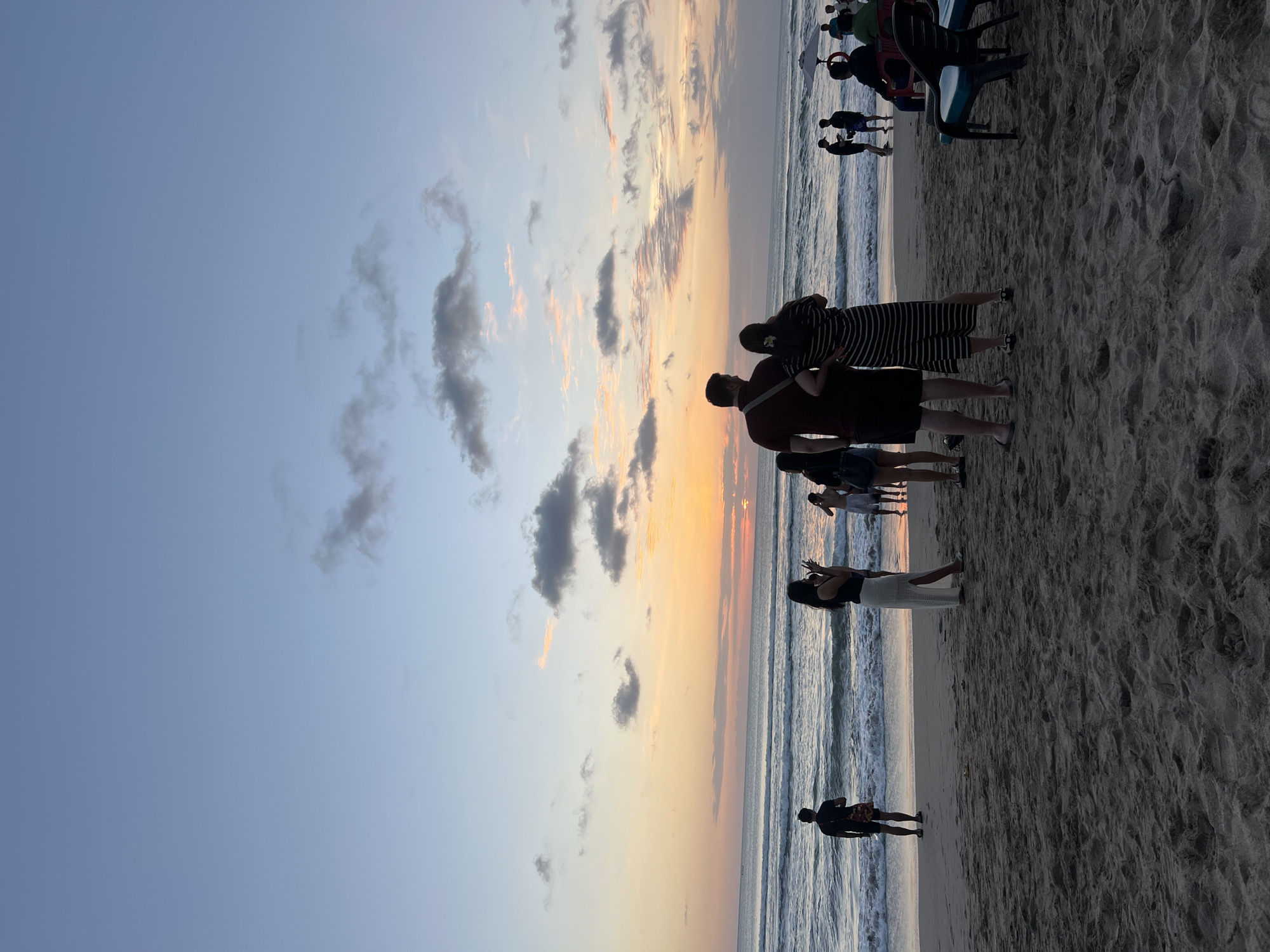 The image depicts a beach scene in Kuta, Indonesia, at dusk. The sky is a gradient of blues, with streaks of orange and pink from the setting sun visible on the horizon. Clouds are scattered across the sky, with some appearing darker and more defined than others. Gentle waves are rolling onto the sandy shore. Several people are present on the beach, some standing near the water's edge, while others are further up on the sand. In the foreground, a couple is standing with their backs to the camera, facing the ocean. Other individuals are scattered throughout the scene, some appearing to be taking photos or simply enjoying the view. Beach chairs are visible on the right side of the image. The overall atmosphere suggests a peaceful evening at a popular tourist destination.