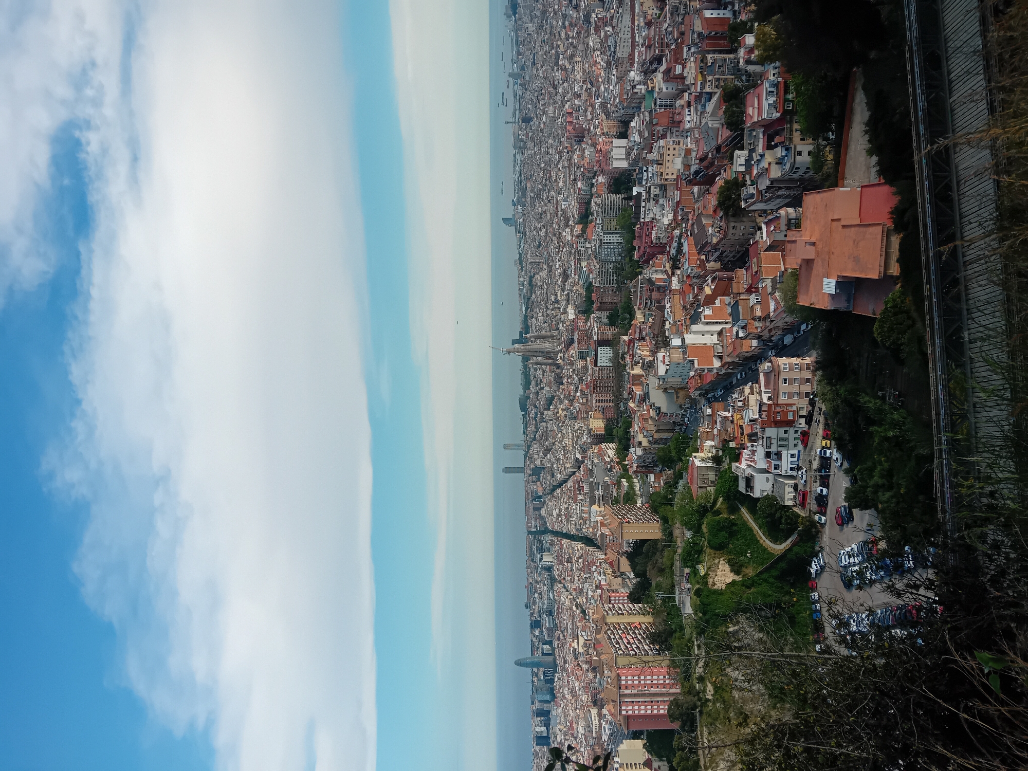 The image offers a high-angle panoramic view of Barcelona, Spain, stretching from a vegetated foreground across a dense urban landscape to the distant Mediterranean Sea, all under a bright, partly cloudy sky. The perspective suggests a viewpoint from a hill or mountain overlooking the sprawling city.
The urban expanse dominates the middle ground, showcasing thousands of tightly packed buildings, predominantly with terracotta-tiled roofs and light-colored facades characteristic of Mediterranean architecture. The iconic Sagrada Familia basilica is clearly visible in the mid-distance, identifiable by its distinctive spires, along with other high-rise buildings and skyscrapers that dot the city's skyline towards the horizon.
In the immediate foreground, dense green foliage and trees partially frame the view, emphasizing the elevated perspective. Below, a winding road leads to a large parking lot filled with numerous cars of various colors. Along the far right edge of the frame, a raised structure resembling a railway track or elevated pathway runs parallel to the cityscape.
The upper portion of the image features a bright blue sky adorned with large, wispy white clouds. The horizon is defined by the calm expanse of the Mediterranean Sea, where a few ships or boats are discernible, appearing slightly hazy due to atmospheric conditions.
The scene depicts a static, daytime view, likely late morning or early afternoon, given the clear visibility and ample sunlight. No specific human activity or event is highlighted. The weather appears clear and pleasant. There is no visible text within the image.