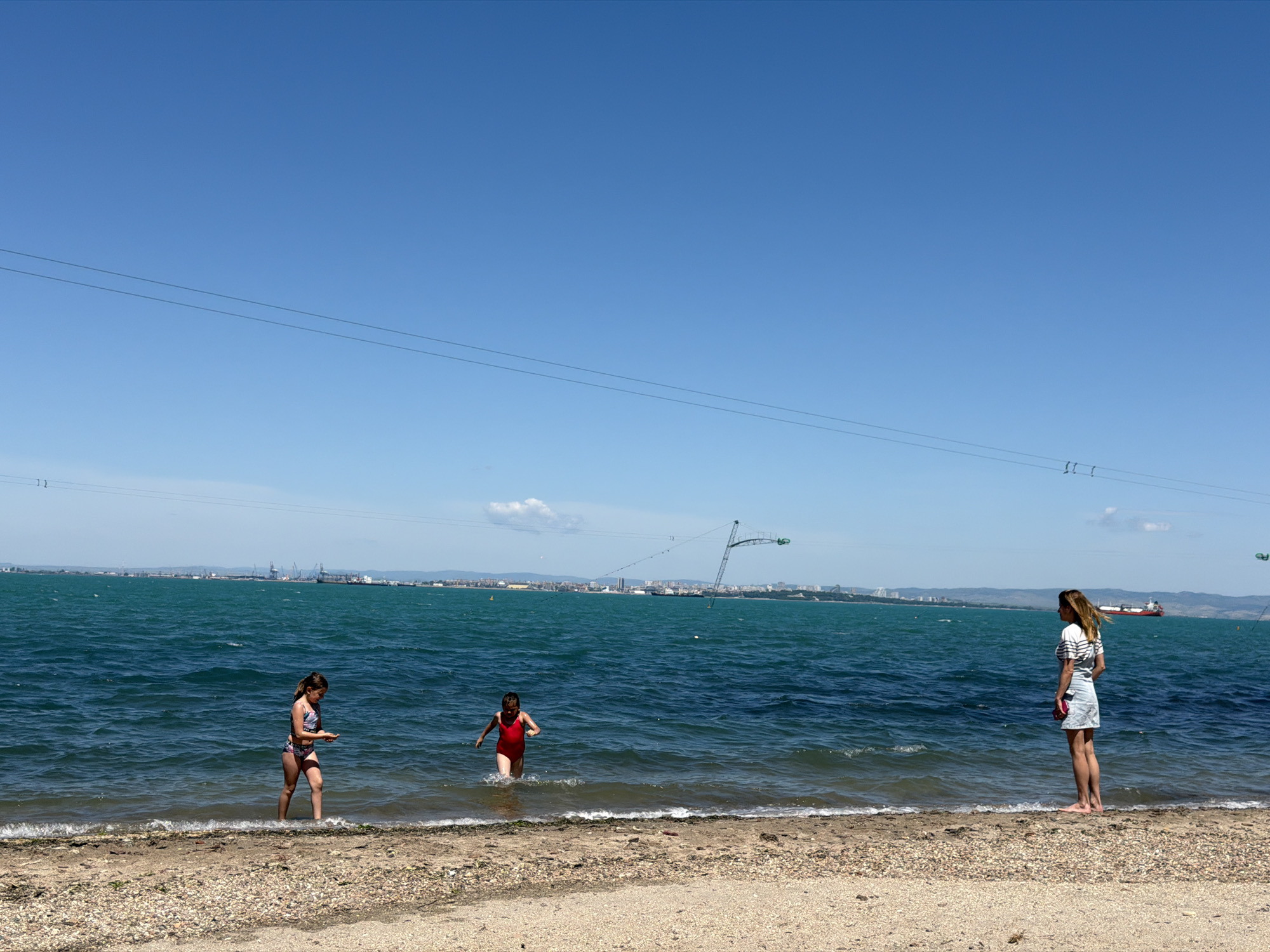 Three individuals are on a beach in Burgas, Bulgaria, on a clear, sunny day. Two young girls, one in a patterned bathing suit and the other in a red one-piece, are wading in the water. A woman stands on the beach's edge, wearing a striped shirt and denim skirt, facing the sea. In the background, the Burgas cityscape is visible, featuring cranes, buildings, and a large cargo ship on the water. Power lines stretch across the clear blue sky. The beach has a sandy surface mixed with small pebbles.