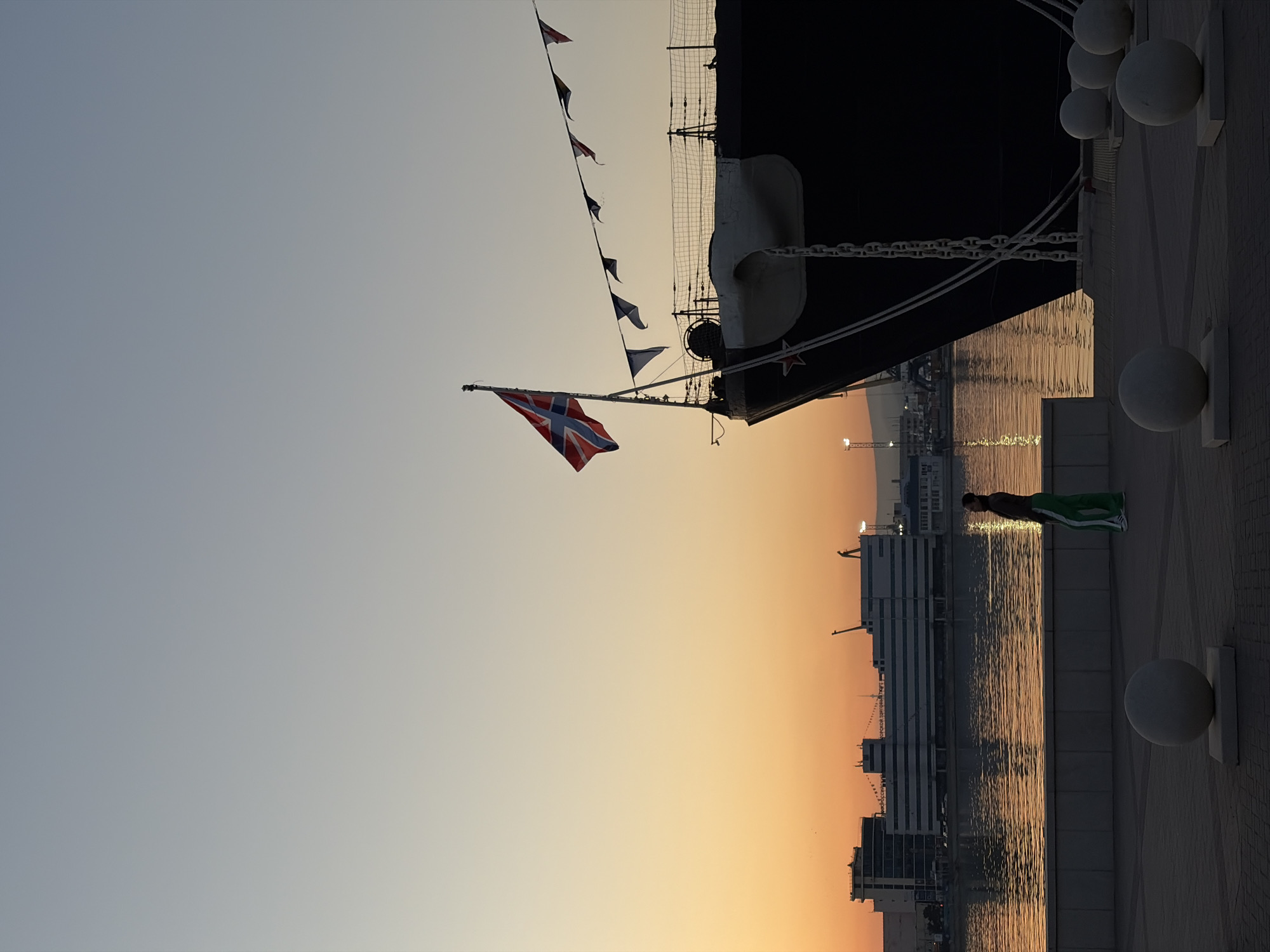 The image captures a partially visible, docked ship with a flag resembling a Union Jack but featuring a navy blue cross instead of red. A string of small flags is also visible. The ship is next to a waterfront promenade, indicated by round bollards. A man wearing a green and white tracksuit stands on the promenade, appearing to be looking at the water. The backdrop includes buildings silhouetted against an orange sunset or sunrise. This is likely taken in Novorossiysk, Russia, given the flag and the visible architecture that aligns with the city's coastal features.