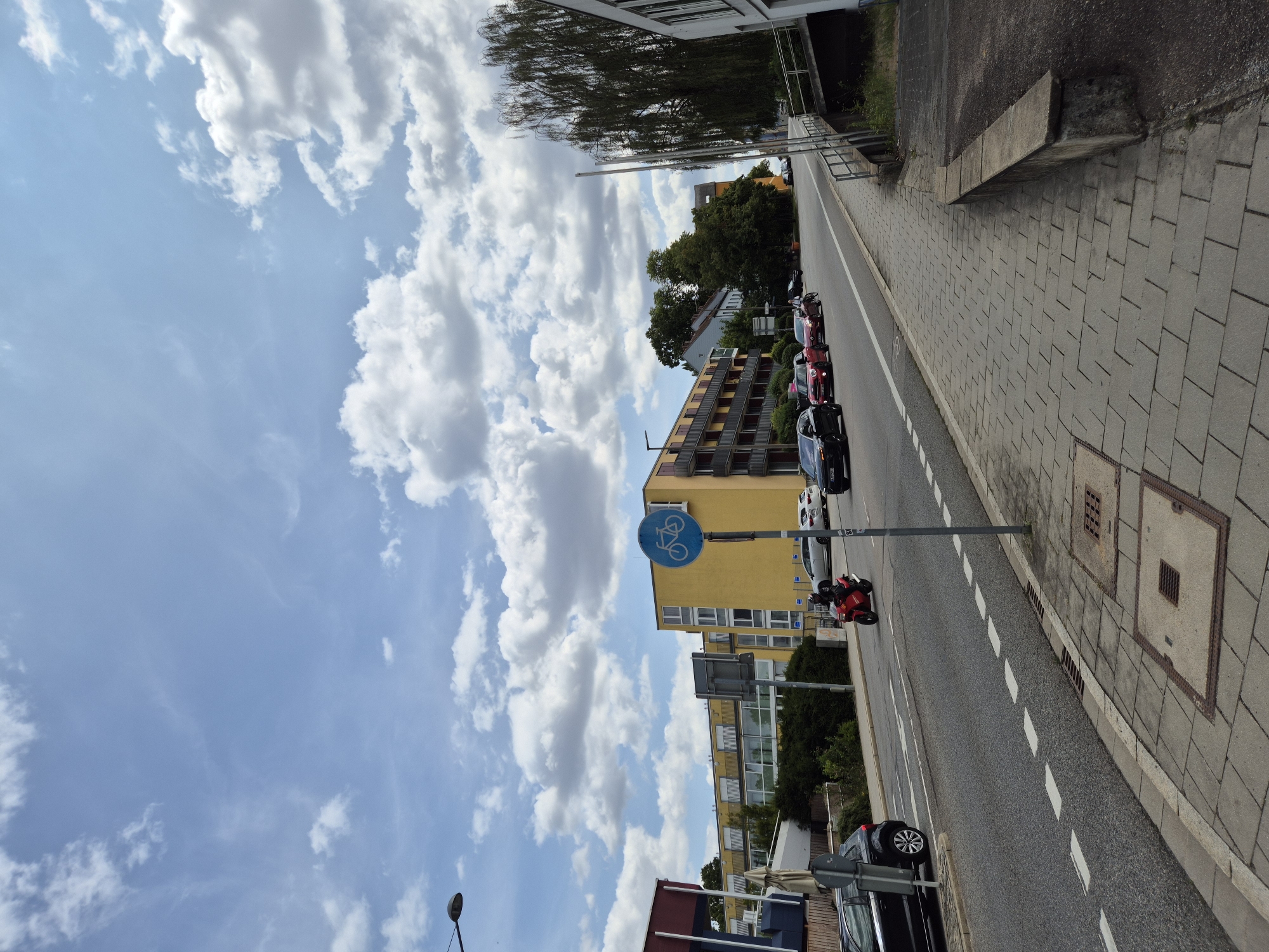 The image captures a street scene in Regensburg, Germany, on a day with a partly cloudy sky. The perspective is from a sidewalk looking towards a street with a moderate incline.
In the foreground, the sidewalk is paved with light grey rectangular stones. A concrete curb separates the sidewalk from the road. Two metal grates are visible on the sidewalk, one with a square opening and the other a larger, rectangular one. A low concrete bench runs along the edge of the sidewalk.
The road is asphalt and shows lane markings, including dashed white lines. Several cars are on the road. From left to right, a black sedan is parked. Further down the road, a red scooter with a rider in black is moving forward. Following the scooter, there are several cars, including a red hatchback, a silver sedan, and other vehicles further in the distance.
On the right side of the road, a grey metal pole supports a blue circular sign with a white bicycle symbol, indicating a bicycle path. Behind the sign, a large, mature weeping willow tree with dense foliage is prominent.
Across the street, a multi-story building with a yellow facade and dark balconies dominates the background. The building has many windows and appears to be residential or commercial. More buildings and trees are visible further down the street, suggesting a suburban or mixed-use area. The sky is a light blue with scattered, fluffy white clouds. A street lamp is visible in the upper left corner of the image.