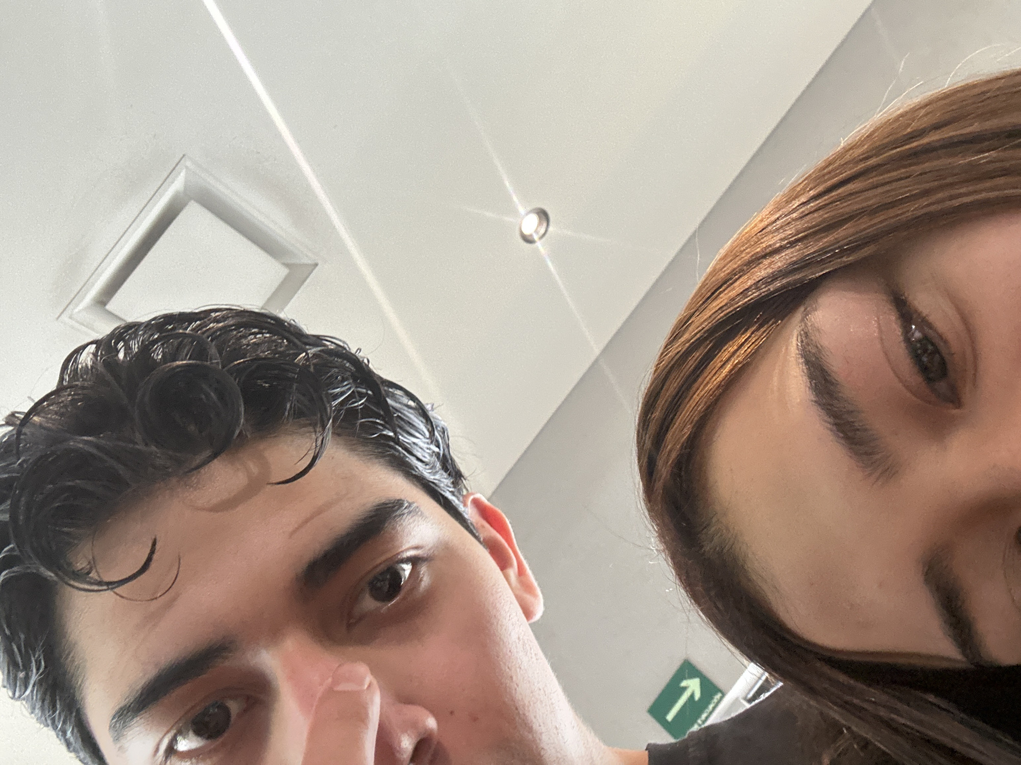 Two individuals are in the frame. The person on the left has short, dark, curly hair, dark eyes, and light skin. Their right index finger is near their nose. The person on the right has long, light-brown hair and light skin. They are both close to the camera, positioned below the lens.
The background shows a white ceiling. There is a square air vent and a recessed light fixture. A green directional sign with a white arrow pointing upwards and to the right is visible near the right individual. Based on the presence of the green directional sign and style of interior, the location is possibly a public space in Puebla, Mexico. However, there are insufficient details to specify the exact location within the city.