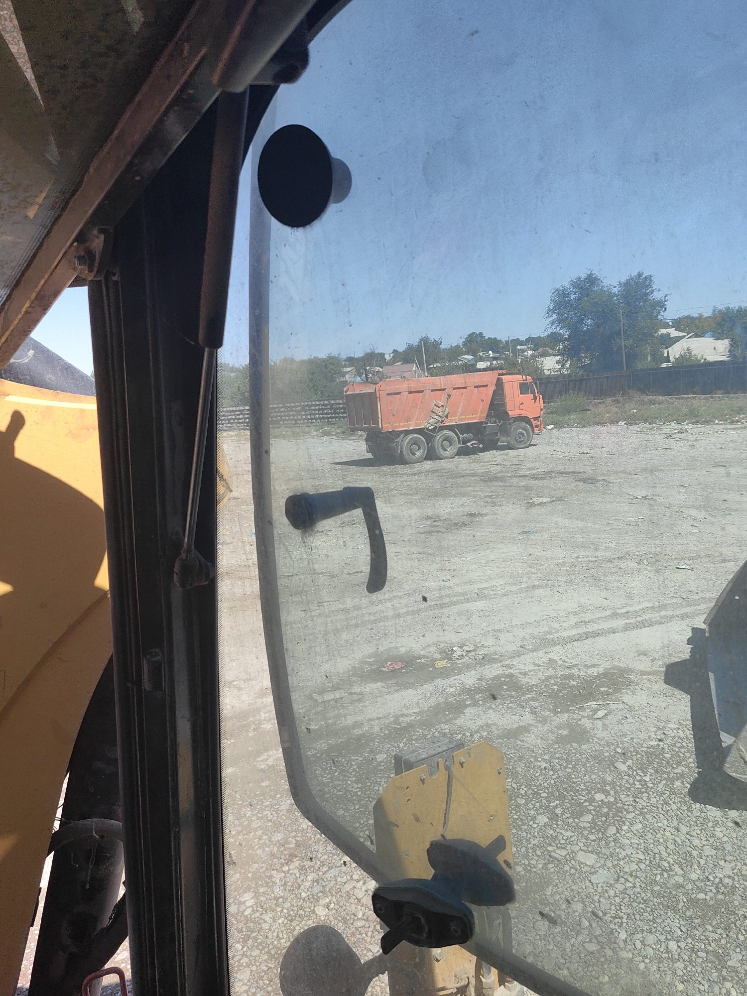 An orange dump truck is parked on a gravel lot. The truck is viewed from inside a cab, looking through a dirty window. In the foreground, a yellow metal piece with two black levers is visible. The cab also contains a black lever mechanism attached to the window frame. The background shows a fence and some buildings, suggesting an industrial or construction area. The sky is clear and blue. The scene appears to be in Shymkent, Kazakhstan.