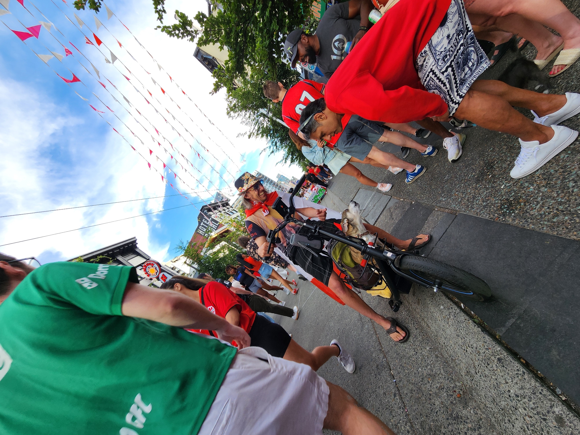 A low-angle, wide shot captures a street scene with a crowd of people dressed in red and green attire, suggesting a sporting event or celebration. In the foreground, a person wearing a green t-shirt with white lettering and white shorts is partially visible on the left. In the center, a man in a red shirt with a bicycle, a dog in the basket, and wearing a patterned helmet is the primary focus. The background shows buildings, trees, and a sky adorned with red and white pennant banners. The overall atmosphere appears festive. The presence of a "HOTEL BENTALL" sign on a building in the background indicates the location is likely in downtown Vancouver, Canada.