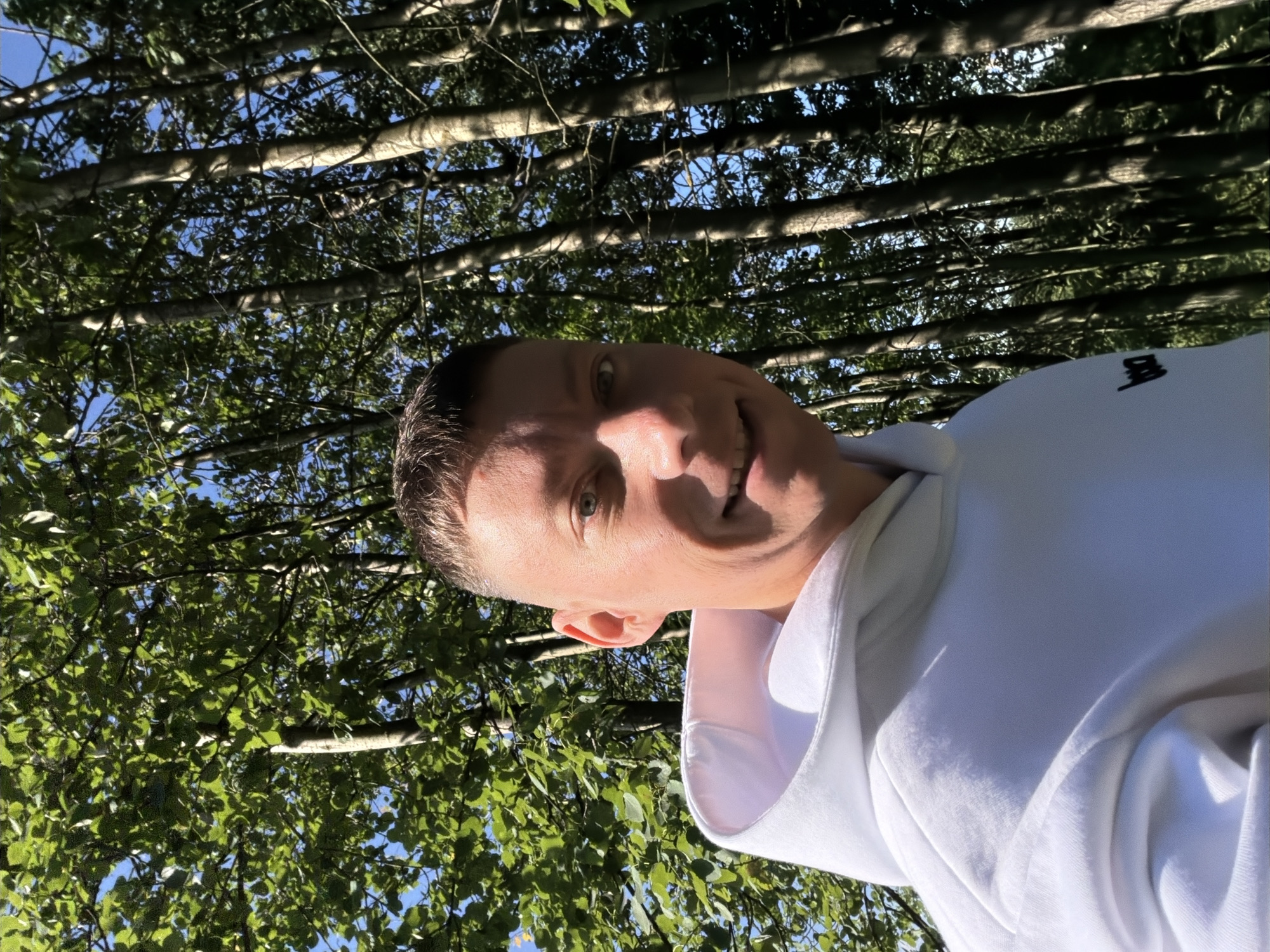 A fair-skinned man with a buzz cut and a white hoodie smiles into the camera, standing outdoors. He is positioned slightly to the right of the frame, with the background dominated by a dense forest of tall, thin trees. The sunlight filters through the leaves, casting dappled shadows. The man's left eye is visible, and his mouth is open in a smile, revealing his teeth. The hoodie has a small black logo on the left side of the chest. There is no indication of the location being Moscow, Russia.