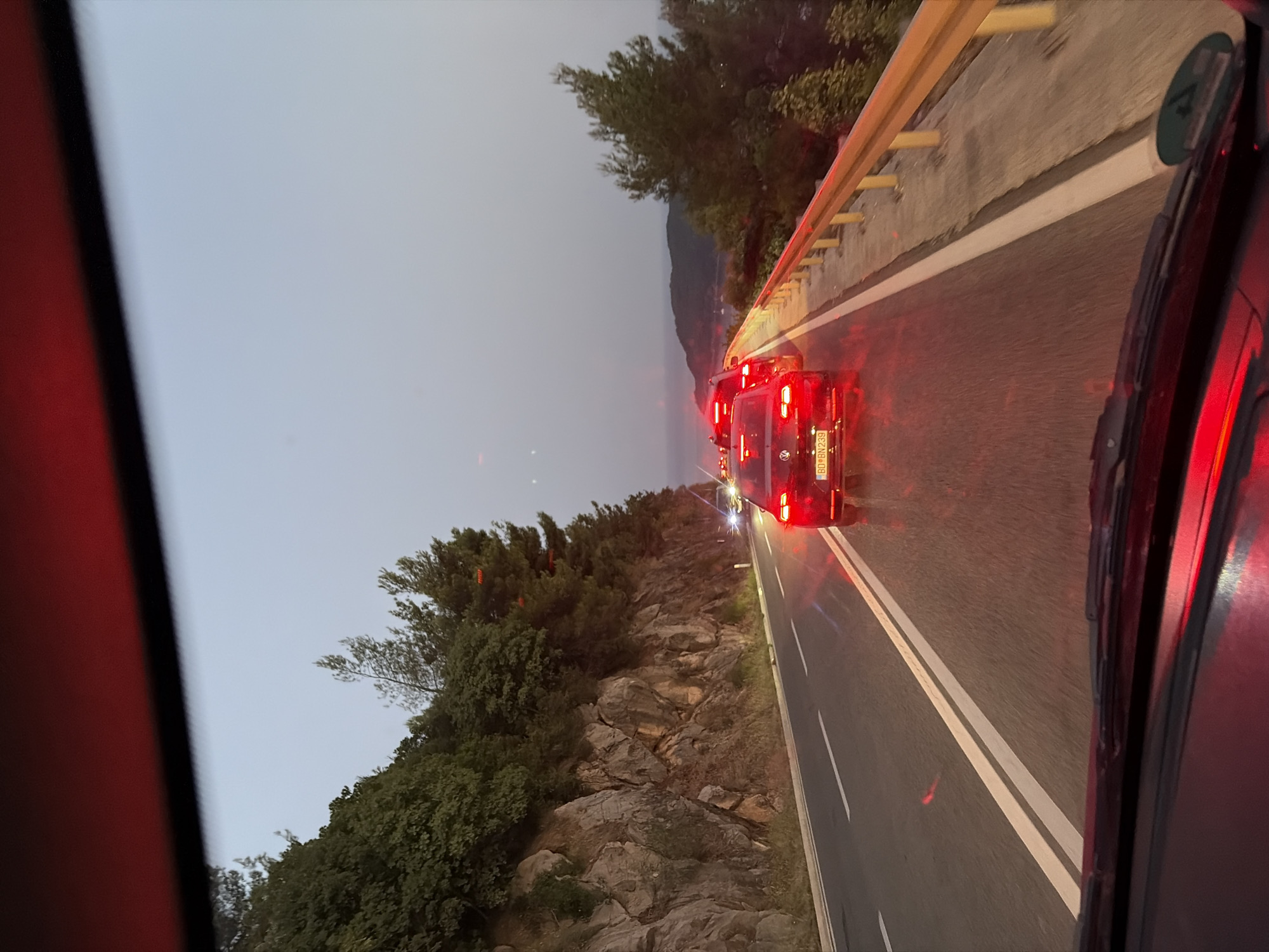 The image shows a view from inside a vehicle traveling on a two-lane road in Košljun, Montenegro. The road is bordered by a steep, rocky embankment on the left, covered in dense green vegetation. On the right, a yellow metal guardrail separates the road from a downward slope with trees. Several cars are visible ahead, appearing to be in a traffic queue. The taillights of these vehicles are illuminated, casting a red glow on the wet road surface. The sky is a pale, hazy gray, suggesting it is either early morning or late evening. In the distance, a body of water and hills are faintly visible, characteristic of the coastal landscape of Montenegro. The foreground shows the dashboard and windshield wipers of the vehicle.