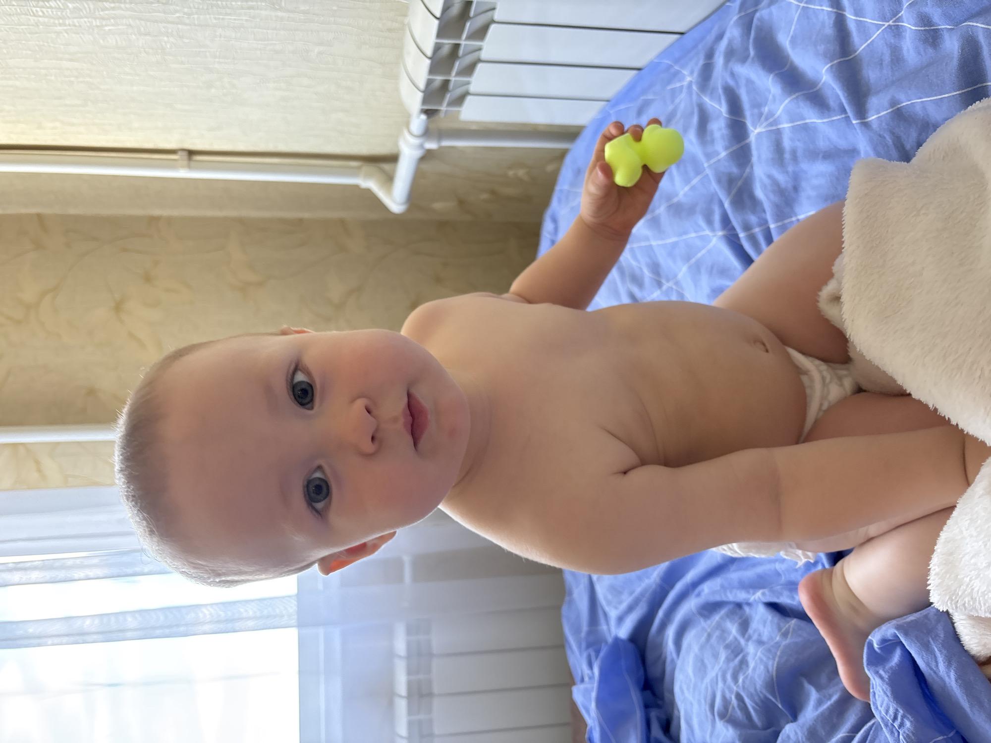 A baby is lying on a blue and white patterned bed. The baby is wearing a diaper and has a green toy in its right hand. The baby has light skin and blue eyes. The background shows a radiator and a window with blinds. The image is taken indoors, possibly in a bedroom. The lighting suggests it could be daytime. The baby appears to be calm or curious. There is no visible text in the image. The location context provided is Naberezhnye Chelny, Russia.