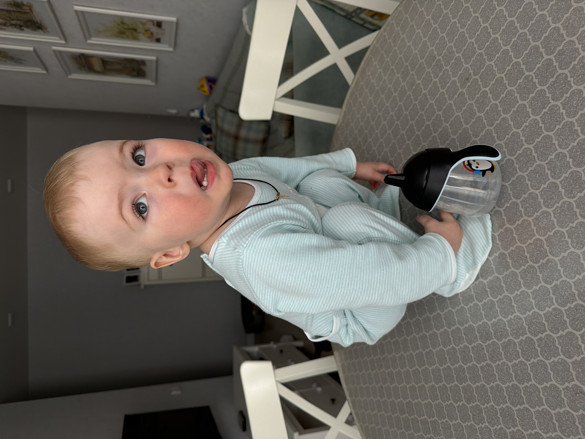 An interior residential scene in Votkinsk, Russia, displays a young child, appearing to be a toddler, squatting centrally on a light grey table surface adorned with a repeating geometric quatrefoil pattern. The child has light blonde hair and blue eyes, is dressed in a light blue and white horizontally striped long-sleeved jumpsuit, and wears a thin black cord necklace with a small gold-colored pendant. The child's gaze is directed upwards and to the right, with the mouth slightly open and tongue partially extended; the right cheek exhibits a mild flush. A clear plastic sippy cup with a black lid, black handles, and a cartoon penguin decal with red headphones is positioned on the table to the child's lower left, adjacent to their left hand. In the background, two white wooden chairs with X-shaped backrests are visible on either side of the table. The room features grey walls, with a black wall-mounted television partially visible to the left and four light-framed pictures hanging on the wall to the right. A light-colored dresser and a sofa with plaid cushions are also discernible in the background, accompanied by various indistinct colorful objects.