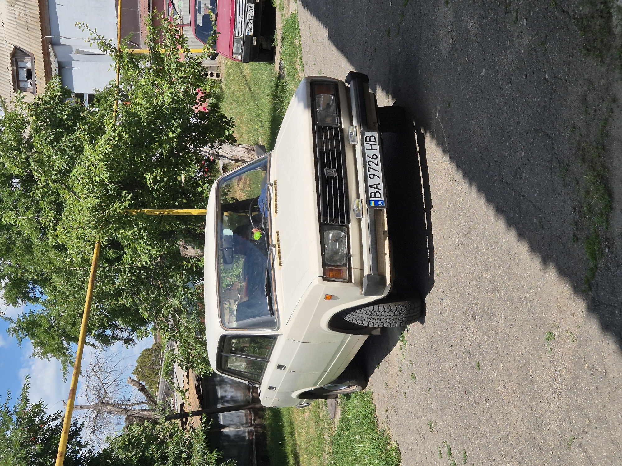 In the village of Sozonivka, Ukraine, on a sunny day, a light-colored sedan, identified by its license plate "BA 9726 HB", is parked on a gravel and asphalt surface. The car is positioned in the foreground, facing slightly to the right. Behind it, a red sedan, with the license plate "AE 2141 AA", is parked. The environment is characterized by green trees and foliage, and a building with a corrugated roof is visible in the background. A yellow pipe runs horizontally above the vehicles and trees. The scene is illuminated by bright sunlight, casting shadows on the ground.