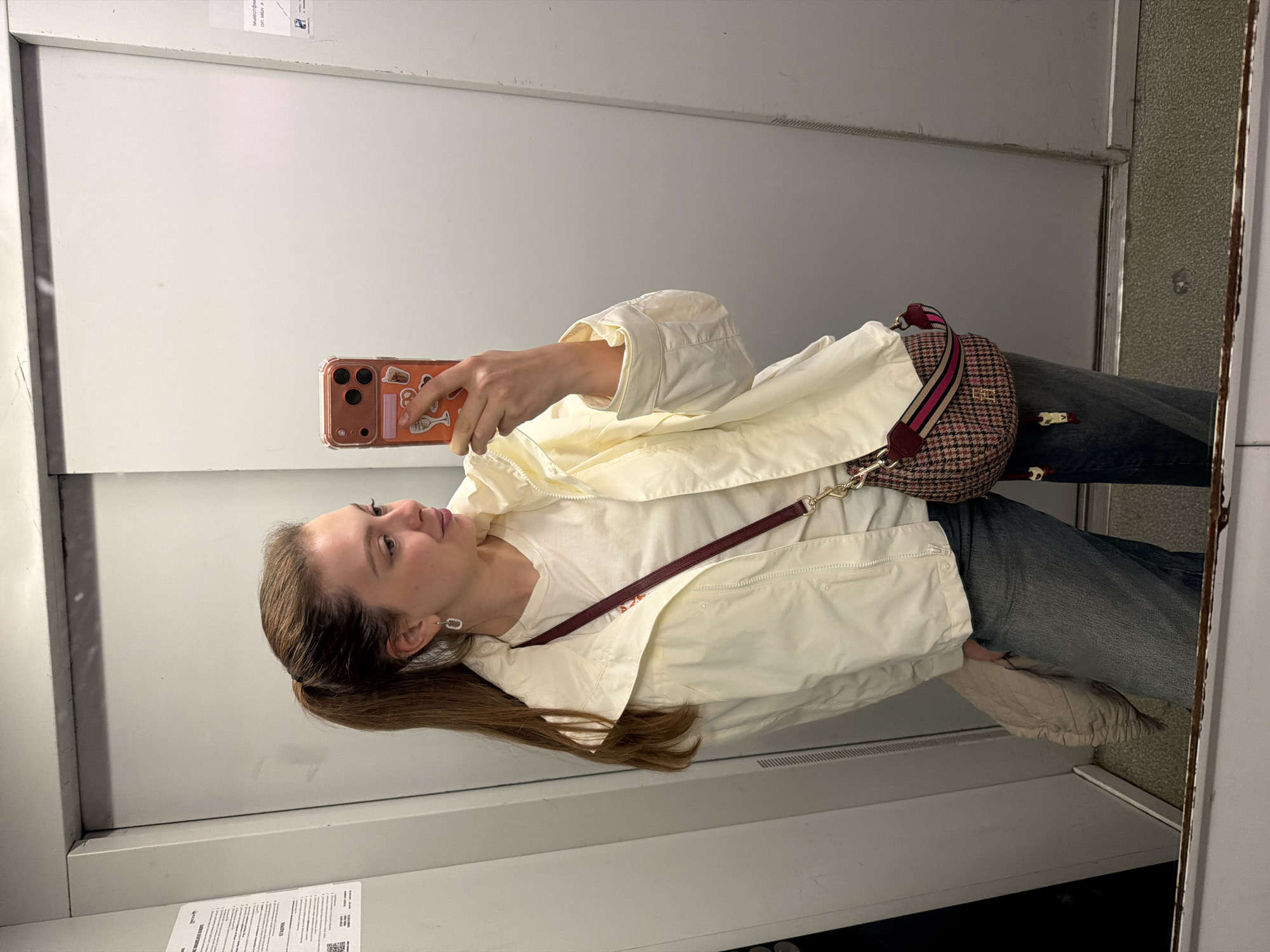 A young woman is taking a selfie in what appears to be a public restroom or elevator, judging by the bland, institutional walls. She is wearing a white t-shirt, a light yellow jacket, and jeans. A brown and pink handbag with a burgundy strap is slung across her body. She is holding a phone with a decorated case. The location of Khimki, Russia, cannot be confirmed based on the visual information provided.