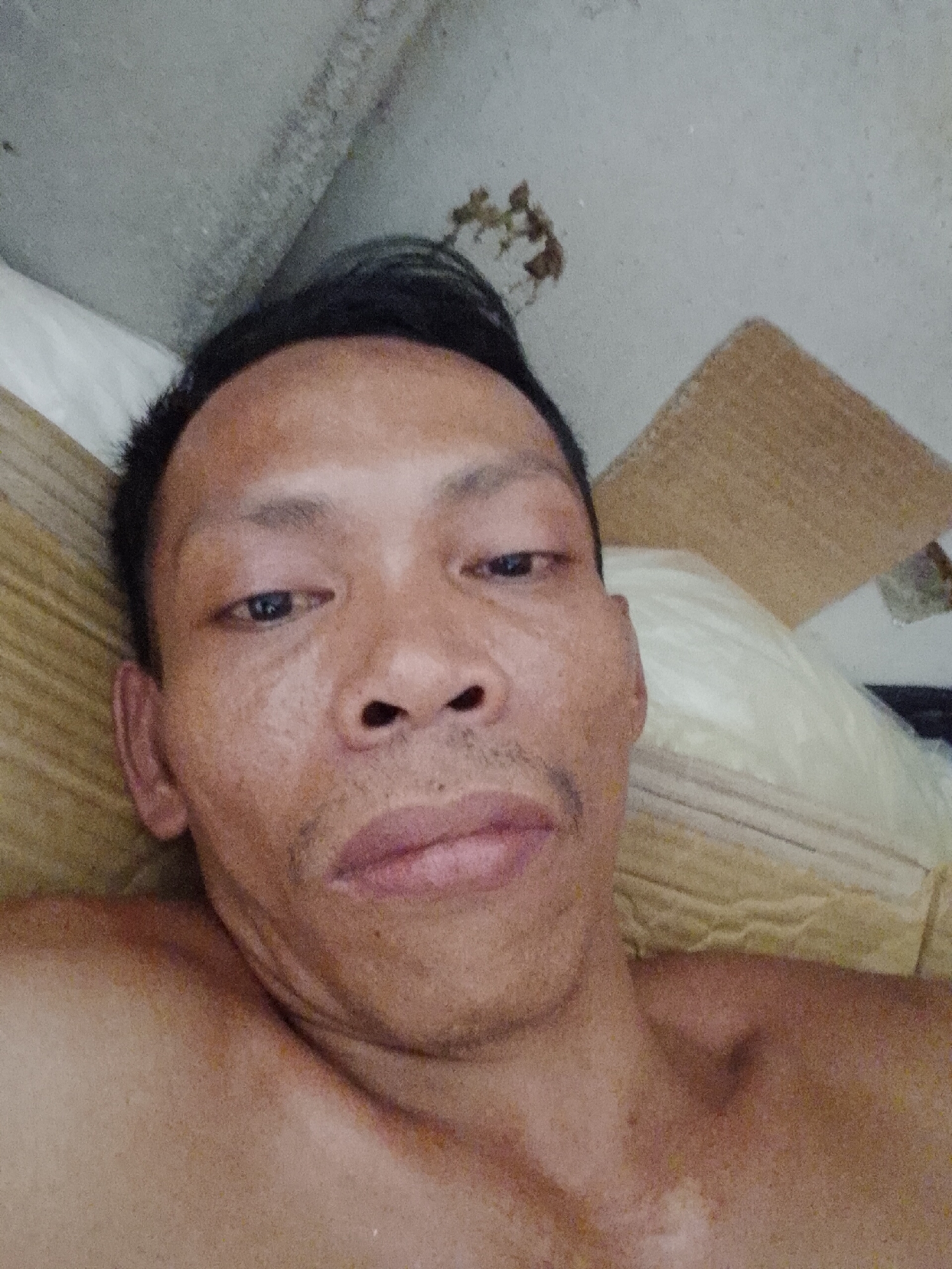 A close-up selfie of an Asian man lying on a tan, textured surface, likely a bed. The man has dark hair and a slight mustache. The background consists of a cream-colored pillow, a cardboard sheet, and a stained ceiling. There is no information to indicate that the picture was taken in the city of Bukit Ketok, Indonesia.