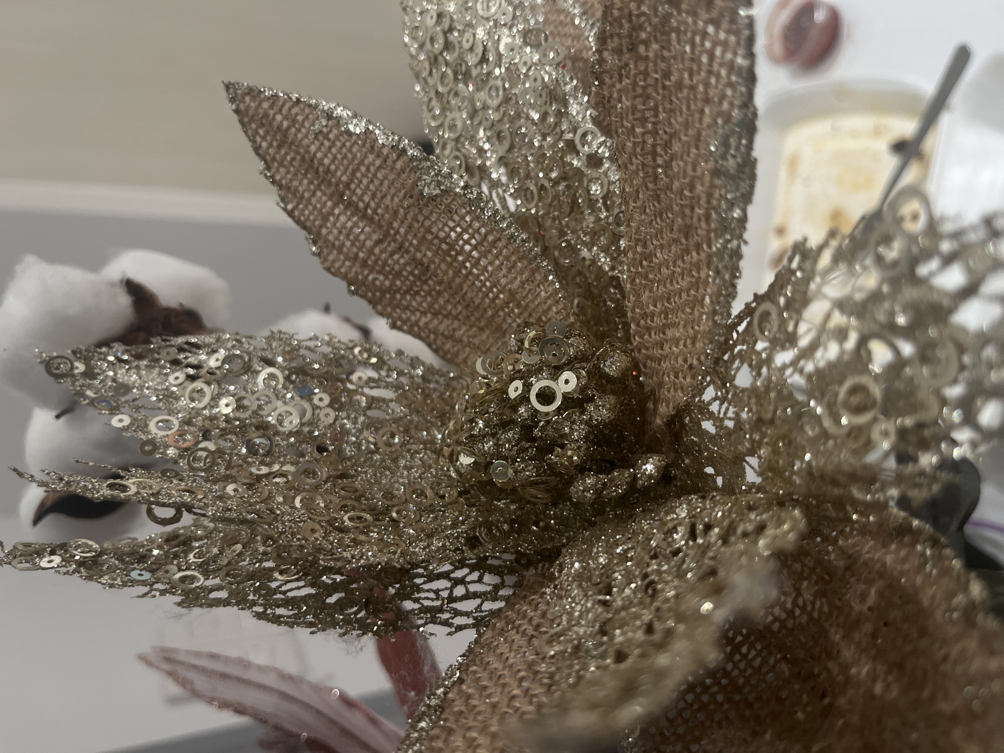 The image presents a close-up view of a large, artificial flower-like decoration. The primary subject is crafted from a textured, golden-brown fabric that resembles burlap, heavily embellished with sparkling gold glitter and numerous small, circular silver sequins. These reflective elements are densely applied across the petals and center of the decoration, creating a shimmering effect. Some areas show a more open, almost lace-like pattern of sequins, while others are solid with glitter.
In the blurred background to the left, several fluffy white cotton bolls are visible, suggesting an autumnal or holiday decorative theme. Further in the out-of-focus background on the right, indistinct objects include what appears to be a white dish or bowl, a slender metallic utensil handle (possibly a fork or spoon), and a small, blurry reddish item.
The scene depicts a static arrangement of decorative items in what appears to be an indoor setting, possibly a home or display area. There is no specific activity taking place, and no people are visible. The lighting is consistent with an indoor environment, but the time of day is not indicated. No visible text is present in the image.