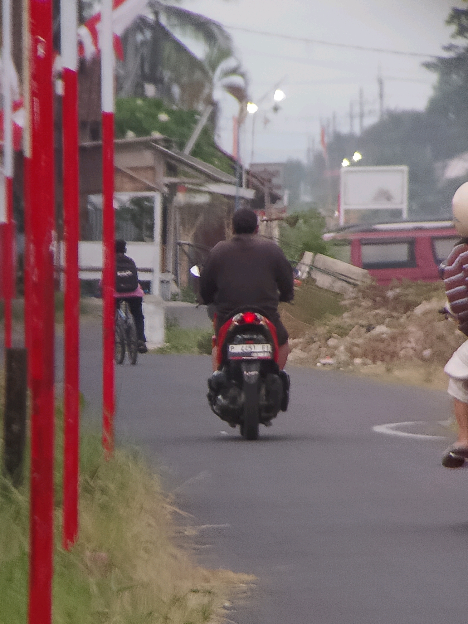 The image depicts a street scene in Jember, Indonesia, characterized by overcast skies and a sense of hurried activity. In the foreground, red and white poles line the left side of the frame, possibly indicating a roadside boundary or barrier. The road surface is asphalt, with some grass and debris visible along the edges.
Two individuals are prominently featured on motorcycles, both heading away from the viewer. The rider in the center is on a dark-colored scooter, wearing a dark jacket and possibly a helmet, with their body angled forward as if accelerating. The rear of the scooter displays a license plate with the inscription "P 4457 KI". To the right, another motorcyclist, partially visible, is wearing a white helmet and a striped shirt. The background reveals a collection of low-lying buildings, some appearing unfinished or in disrepair, with scattered rocks and foliage. A red vehicle is parked or moving in the mid-ground. Overhead, power lines and streetlights are visible, suggesting an urban or semi-urban setting. The overall atmosphere is somewhat muted, likely due to the cloudy weather.