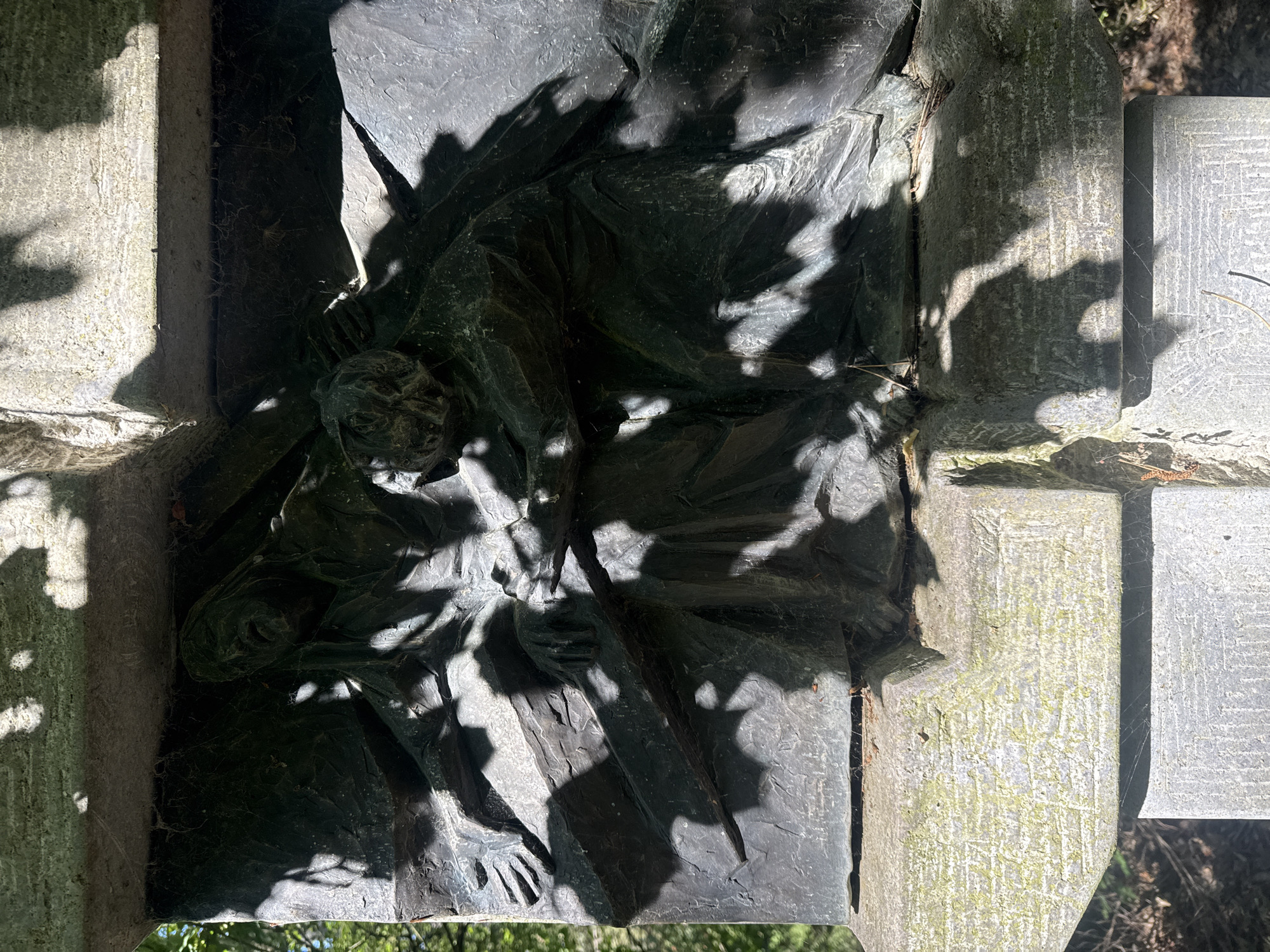 This is a close-up, eye-level photograph of a dark metal bas-relief sculpture depicting a scene with multiple figures. The sculpture is mounted on a stone structure, part of which is visible at the bottom and top of the frame. The bas-relief shows at least two figures in distress, one appearing to be carrying a burden or being helped. Their faces are contorted in anguish, and their hands are reaching out. The sculpture is rendered with dramatic chiaroscuro due to strong sunlight casting dappled shadows from overhead foliage. Cobwebs are visible across the surface of the sculpture, indicating it is outdoors and likely not frequently cleaned.
The stone structure beneath the sculpture is roughly hewn and weathered, with some areas showing greenlichen growth. Faint, incised markings are present on the stone, though they are difficult to discern due to the texture and lighting. The overall impression is one of a somber monument or memorial, likely situated in a cemetery or a park.
The bas-relief is part of the central monument in the war memorial of Bad Salzschlirf, Germany, located in the town's cemetery. The monument is dedicated to the fallen soldiers of both World Wars. This specific scene is believed to represent the suffering and hardship endured by civilians during wartime.