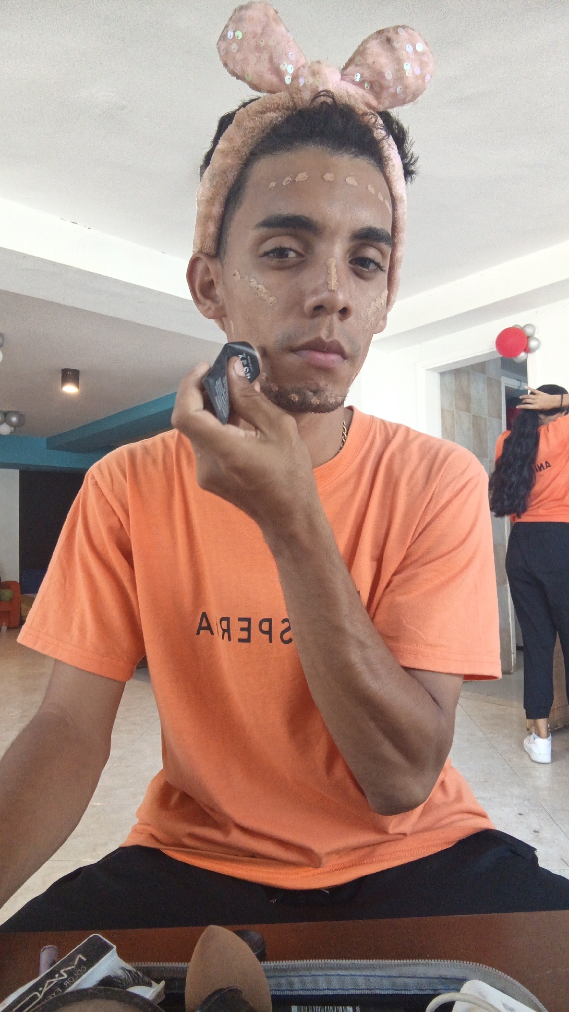 A young adult male is applying makeup in an interior space. He is wearing a peach-colored headband with bunny ears, an orange t-shirt with a mirrored inscription "SPERA," and black pants. Spots of foundation are visible on his face, and he is using a black makeup applicator to blend the makeup on his cheek.
In the background, a woman with long, black hair is wearing a matching orange t-shirt and black pants. A red balloon is visible near the doorway. The location appears to be an interior room with white walls and a teal accent wall. Based on the overall scene and the reference provided, the location is in the city of Playa El Agua, Venezuela.