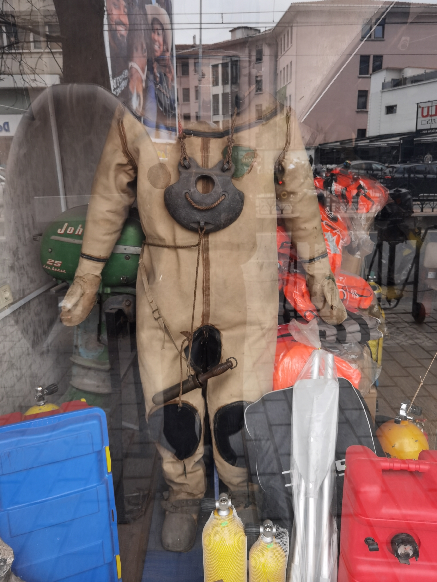 This media file captures a shop window display in Beyoğlu, Turkey, filled with marine and diving equipment, heavily overlaid with reflections of the bustling street scene outside.
The primary subject inside the shop window is a mannequin dressed in an old-fashioned, beige or tan heavy-duty diving suit, possibly a dry suit, complete with attached gloves and boots. A dark, weighted chest piece or collar, possibly made of metal, hangs from its neck, featuring a rope resembling a smile. A knife or tool is strapped to the right leg of the suit.
Behind the mannequin, a vintage-style green outboard motor is visible, with "John" and "25 horsepower" written on its side, likely a Johnson brand engine. Around the display, various marine supplies are arranged, including stacks of bright orange life jackets, small yellow scuba tanks with mesh covers, and distinct red and blue plastic containers, possibly fuel tanks or storage boxes. A set of silver paddles is also part of the display.
The shop window strongly reflects the outdoor environment. Reflections show parts of buildings with a mix of architectural styles, including a prominent light pink building, indicative of the urban setting. Bare tree branches, power lines, and several cars on a street are also visible in the reflections. Additionally, a large poster or advertisement is reflected, showing two smiling individuals, one appearing to wear a cowboy hat, adding a human element to the otherwise inanimate reflections.
The overall impression is that of an overcast day, with diffused daylight illuminating the scene. The multiple layers of reflections create a complex visual, blending the items on display with the street life of Beyoğlu.