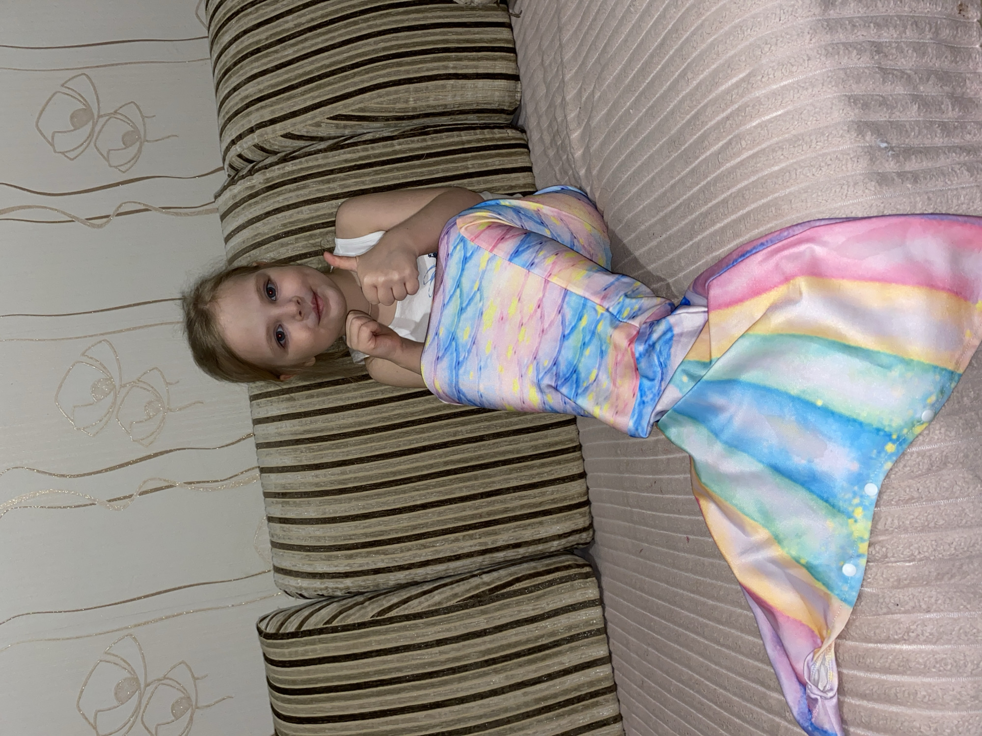 A young girl is captured lying on her side on a striped couch in an indoor setting, likely a home in Votkinsk, Russia. She has blonde hair and blue eyes and is looking directly at the camera with a bright smile. Both of her hands are raised, giving a thumbs-up gesture, indicating happiness or approval.
The girl is dressed in a white tank top, and her lower body is encased in a vibrant mermaid tail costume. The costume features a blend of pastel colors, including pink, blue, yellow, purple, white, and green, adorned with a subtle scale-like pattern and scattered colorful speckles.
The couch she rests on has prominent striped cushions, featuring alternating bands of brown, beige, and dark brown. Behind her, a wall is visible, covered in light-colored wallpaper. This wallpaper is decorated with elegant, stylized floral or abstract designs in a light brown or gold hue, accentuated with glitter details. The lighting suggests an indoor environment, possibly during the day or under artificial light in the evening. There is no visible text in the image. The overall scene conveys a cheerful and playful mood.