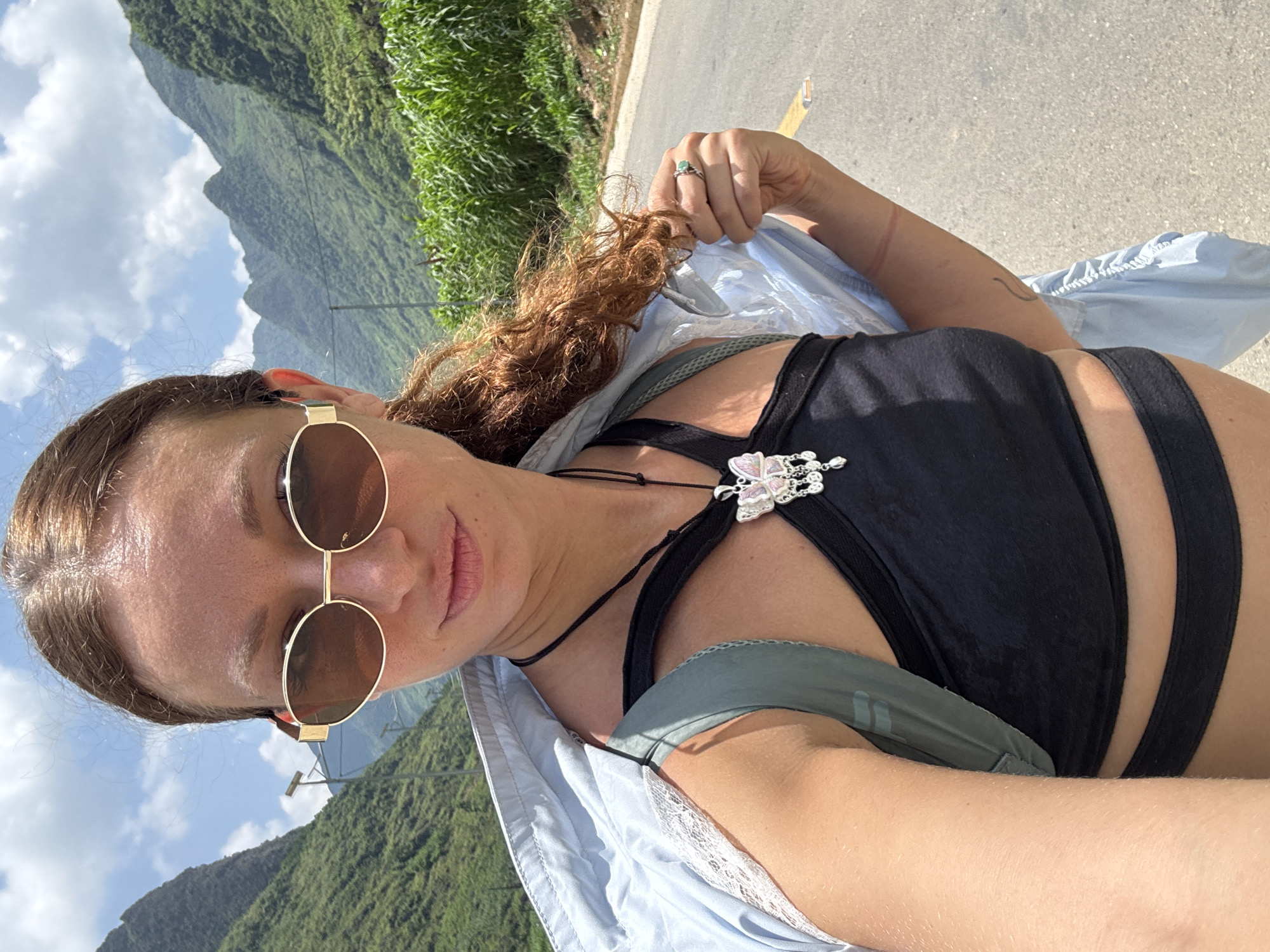 A woman is photographed at the Mèo Vạc Commune in Vietnam. The woman is wearing round sunglasses with a gold frame, a black crop top, and a light blue jacket with a lace trim. She has brown hair and is wearing a ring on her finger and a necklace with a butterfly pendant. In the background are green mountains and a partly cloudy sky. She stands on a gray road with a yellow stripe.